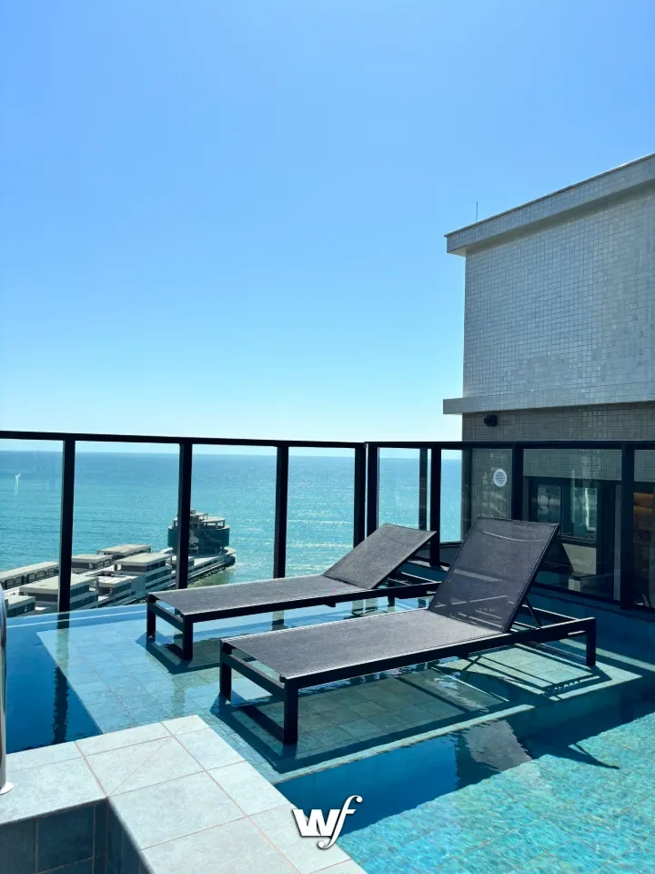 Varanda de piscina com duas espreguiçadeiras deitada sobre o piso de azulejo azul, com visão para o mar e céu claro, ao lado de um prédio de cor cinza.