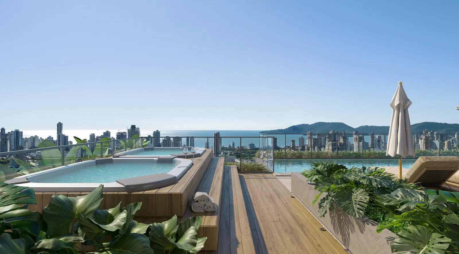 Piscina de borda infinita com vista para a cidade e o mar ao fundo, espreguiçadeiras, guarda-sol branco, plantas verdes, toalhas enroladas, céu claro e ensolarado.