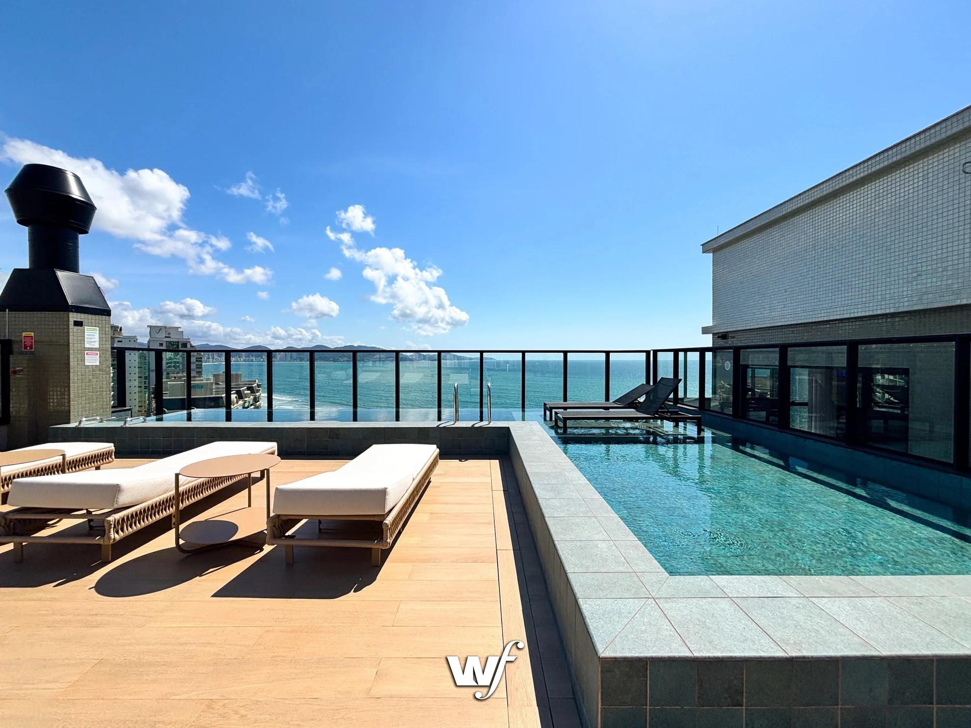 Piscina com espreguiçadeiras em um terraço com vista para o mar azul, céu claro e algumas nuvens, em um edifício alto.
