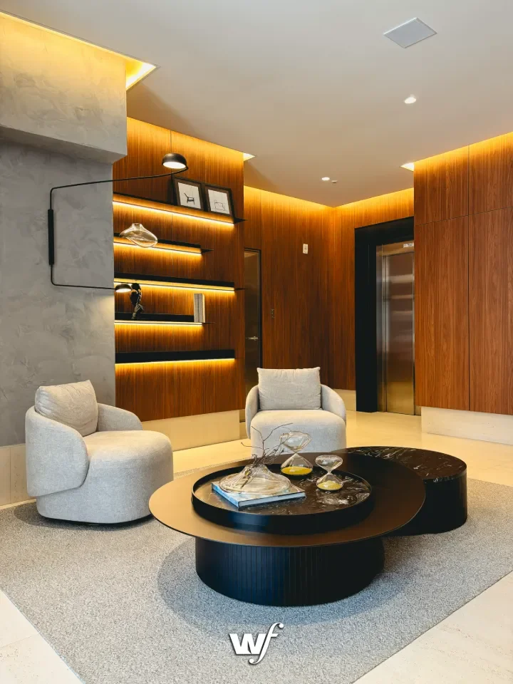 Sala de estar moderna com duas poltronas brancas, mesa de centro preta com objetos decorativos e livros, paredes de madeira, iluminação embutida e detalhes de design minimalista.