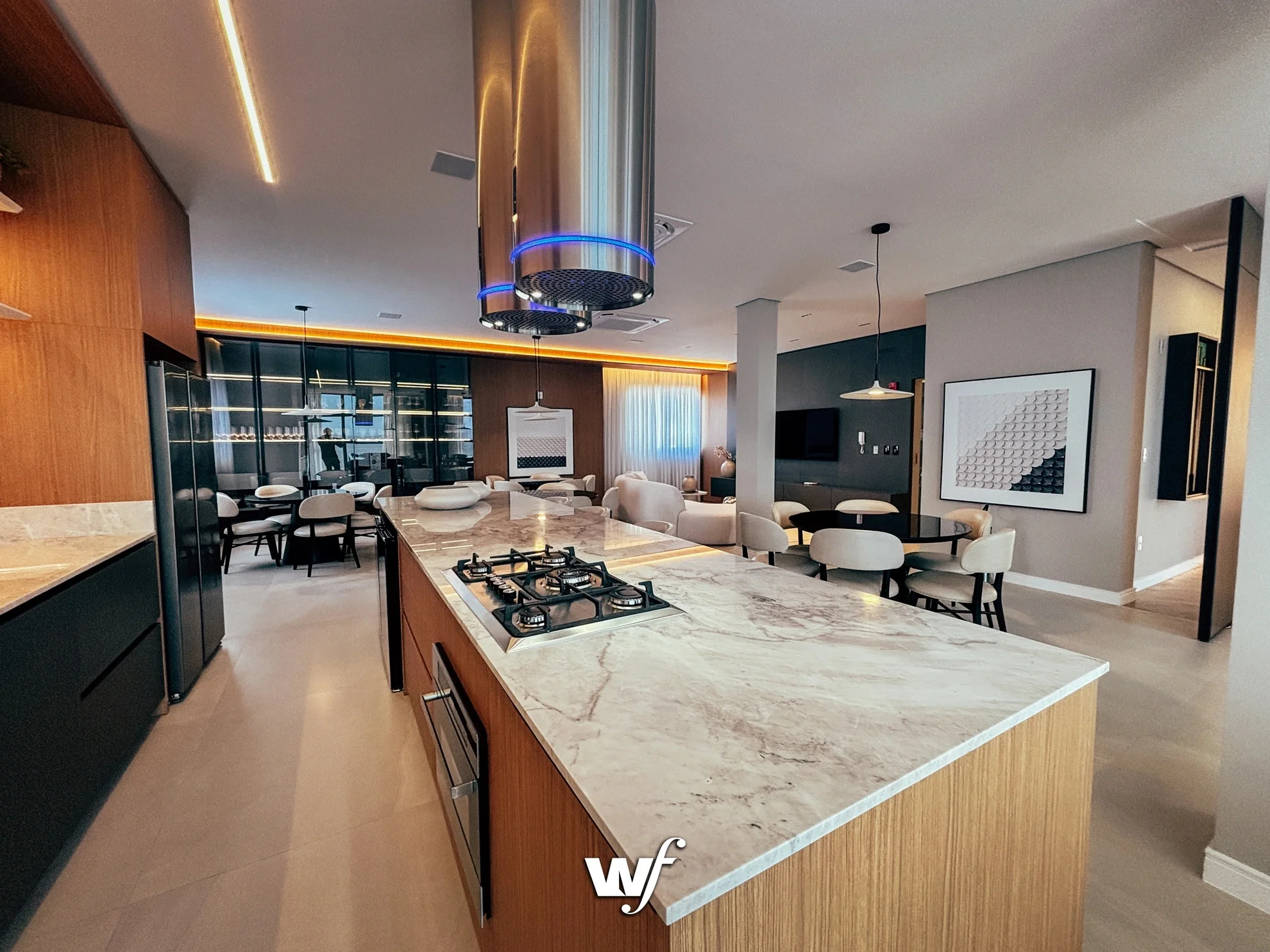 Imagem de uma cozinha moderna com ilha central de granito, cooktop e coifa de inox suspensa. Ao fundo, há uma sala de jantar com mesa redonda e cadeiras brancas, além de uma área de estar com televisão e quadros decorativos nas paredes.