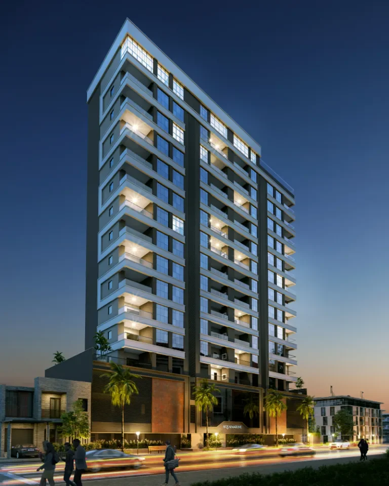 Imagem de um prédio alto de vários andares à noite, com iluminação nas varandas e janelas, rodeado por árvores e uma rua movimentada com carros em movimento.