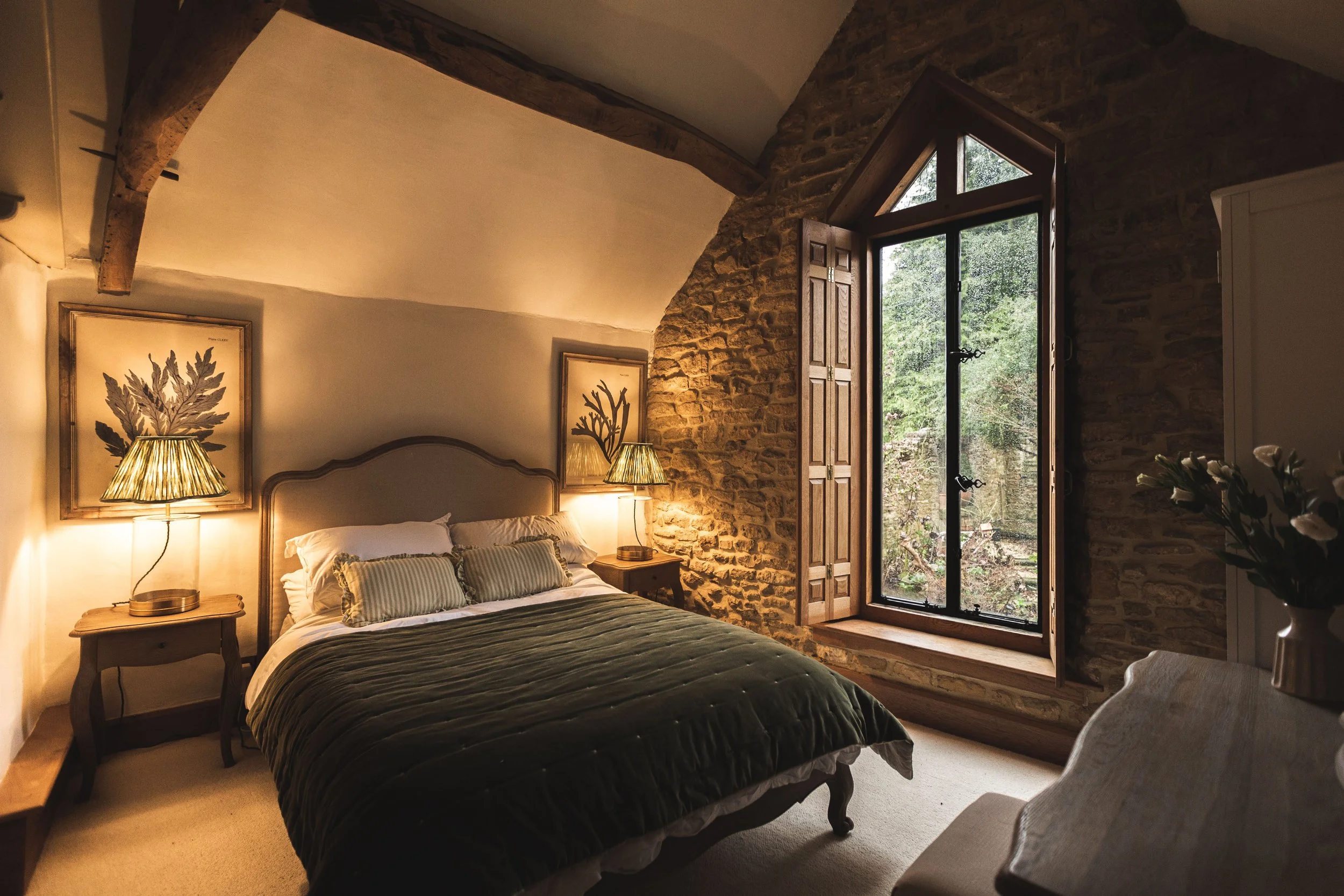Cozy bedroom with a large bed, pillow, and green bedding, illuminated by table lamps. The room features a rustic stone wall, a wooden window with open shutters showing greenery outside, and framed botanical artwork above the bed.