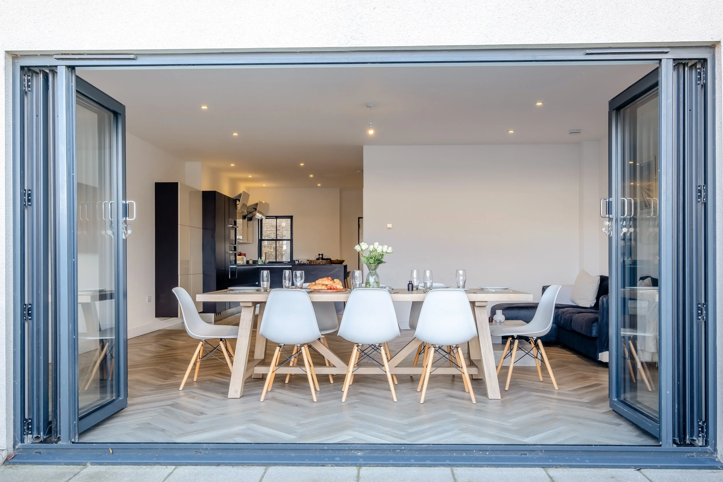 Open foldable glass doors leading into a modern dining room with a wooden table set with glasses and a vase of white flowers, adjacent to a black sofa.