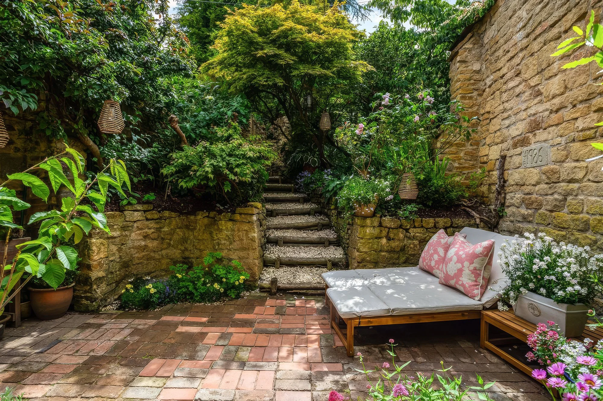 A cozy outdoor patio with a white cushioned lounge chair with pink floral pillows, surrounded by lush green plants, potted flowers, and a stone staircase leading up a garden. A brick wall and garden lanterns are also visible.