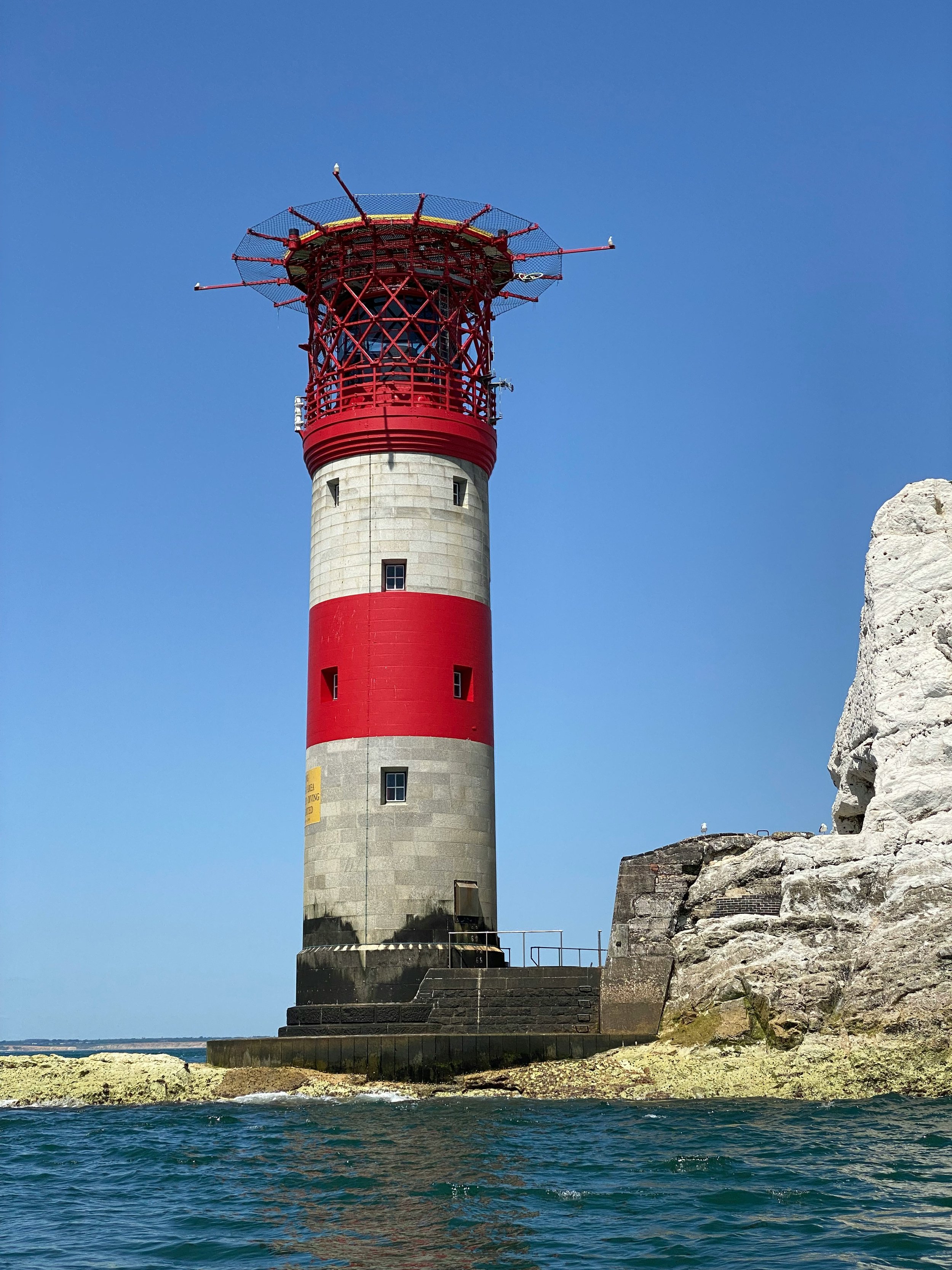 A lighthouse with a gray, red, and white striped tower, located on a rocky shoreline, with a clear blue sky background.