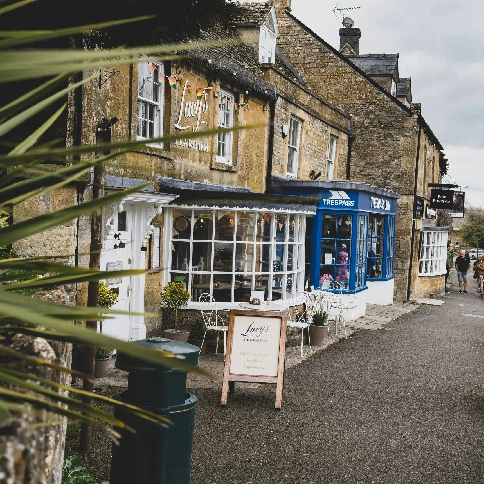 Stone building with storefronts, including Lucy's Tearoom with outdoor seating, a signboard, and a blue Trespass store, on a cloudy day with pedestrians in the background.