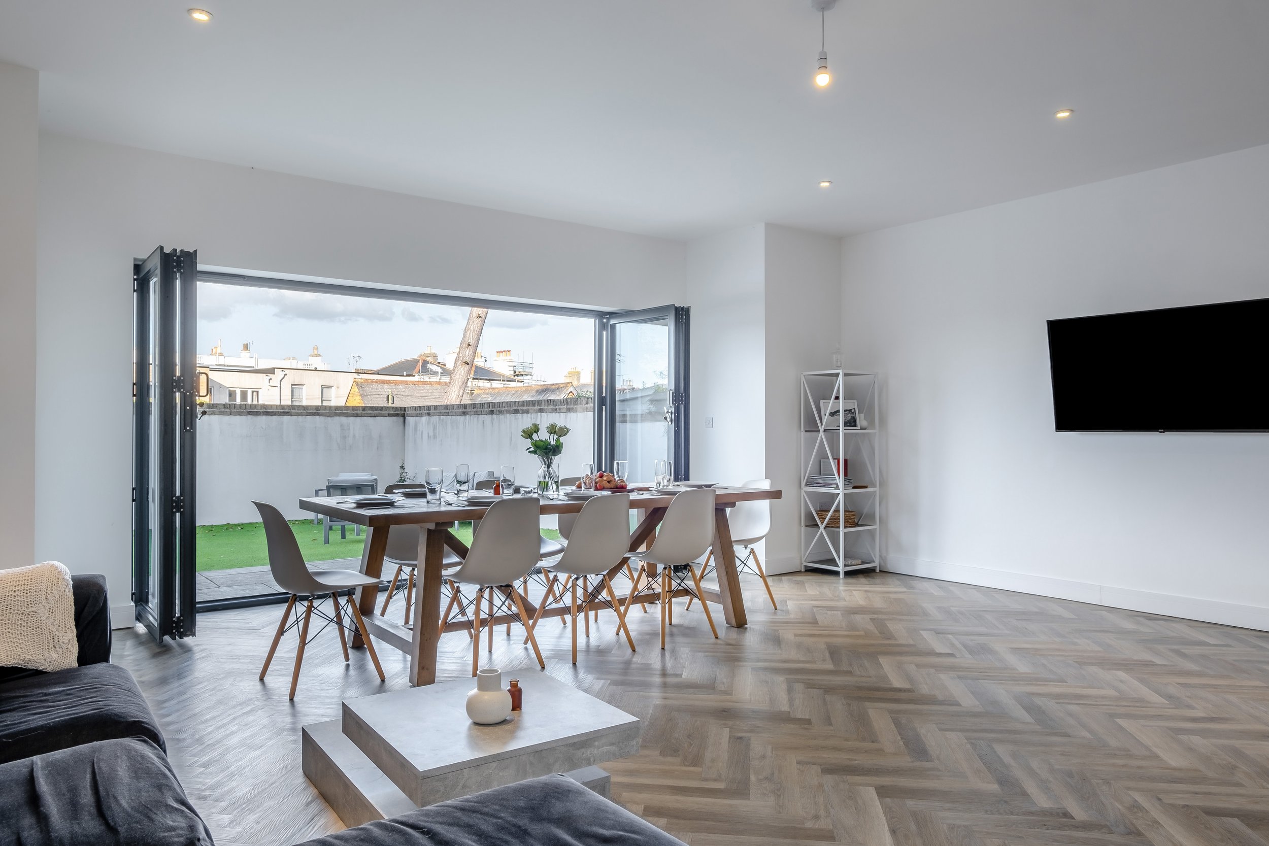 Modern living room with open large window sliding doors, wooden dining table with six white chairs, a small white shelf with decor, a wall-mounted TV, and a grey sofa in the foreground. The room has light-colored hardwood floors and minimal decor.