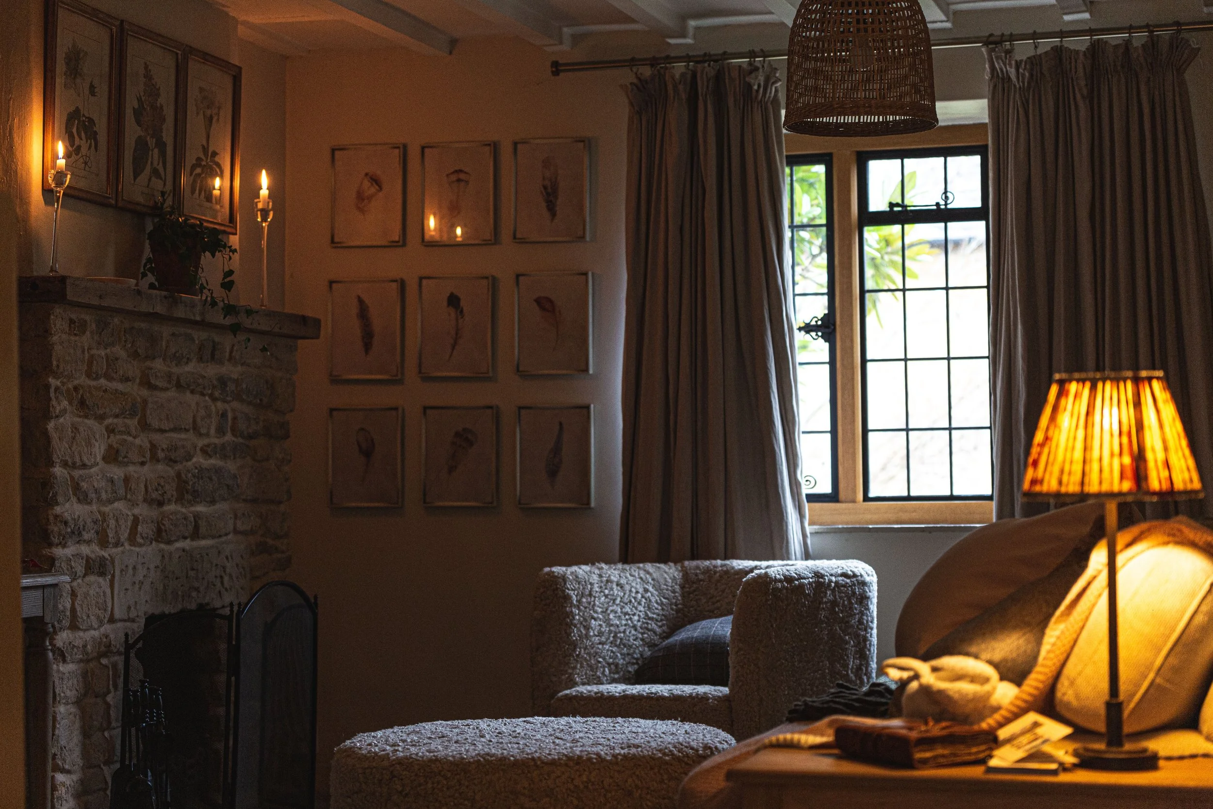 Cozy living room with a stone fireplace, framed botanical prints on the wall, curtains, a window showing greenery outside, a cushioned armchair, a side table with a lamp, and lit candles and lamps creating a warm atmosphere.
