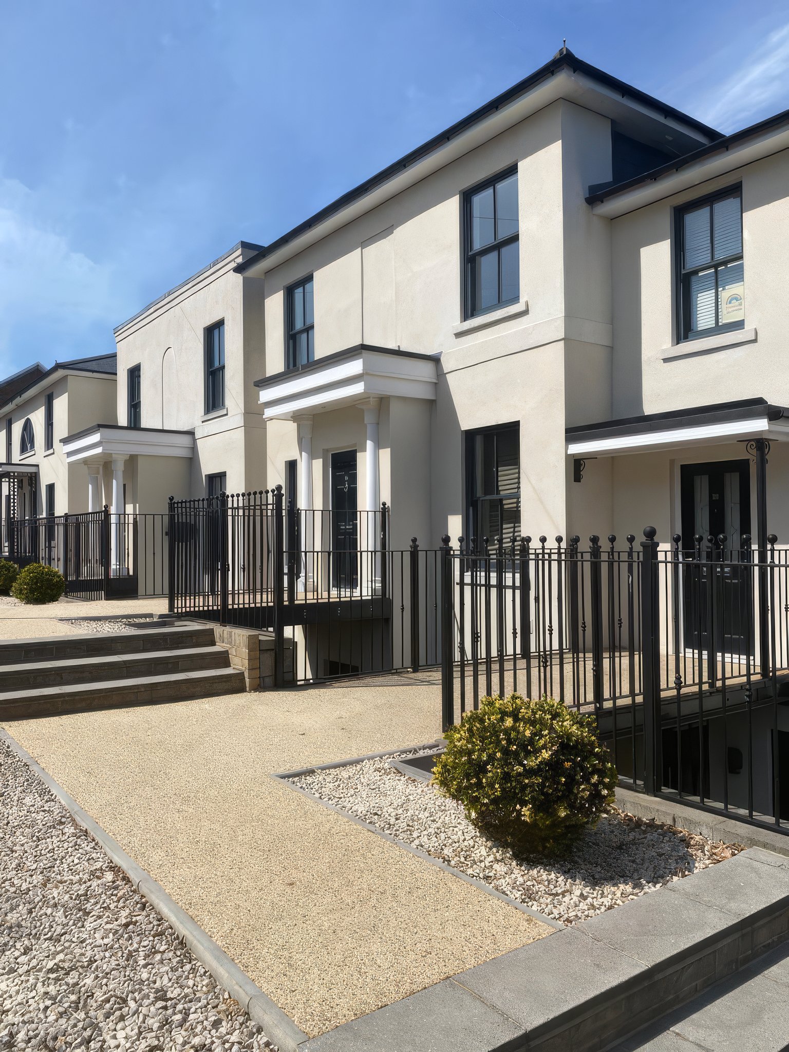 Modern row of white townhouses with black doors and metal railings, small landscaped front yard with a bush and steps leading to the entrance, under a blue sky.