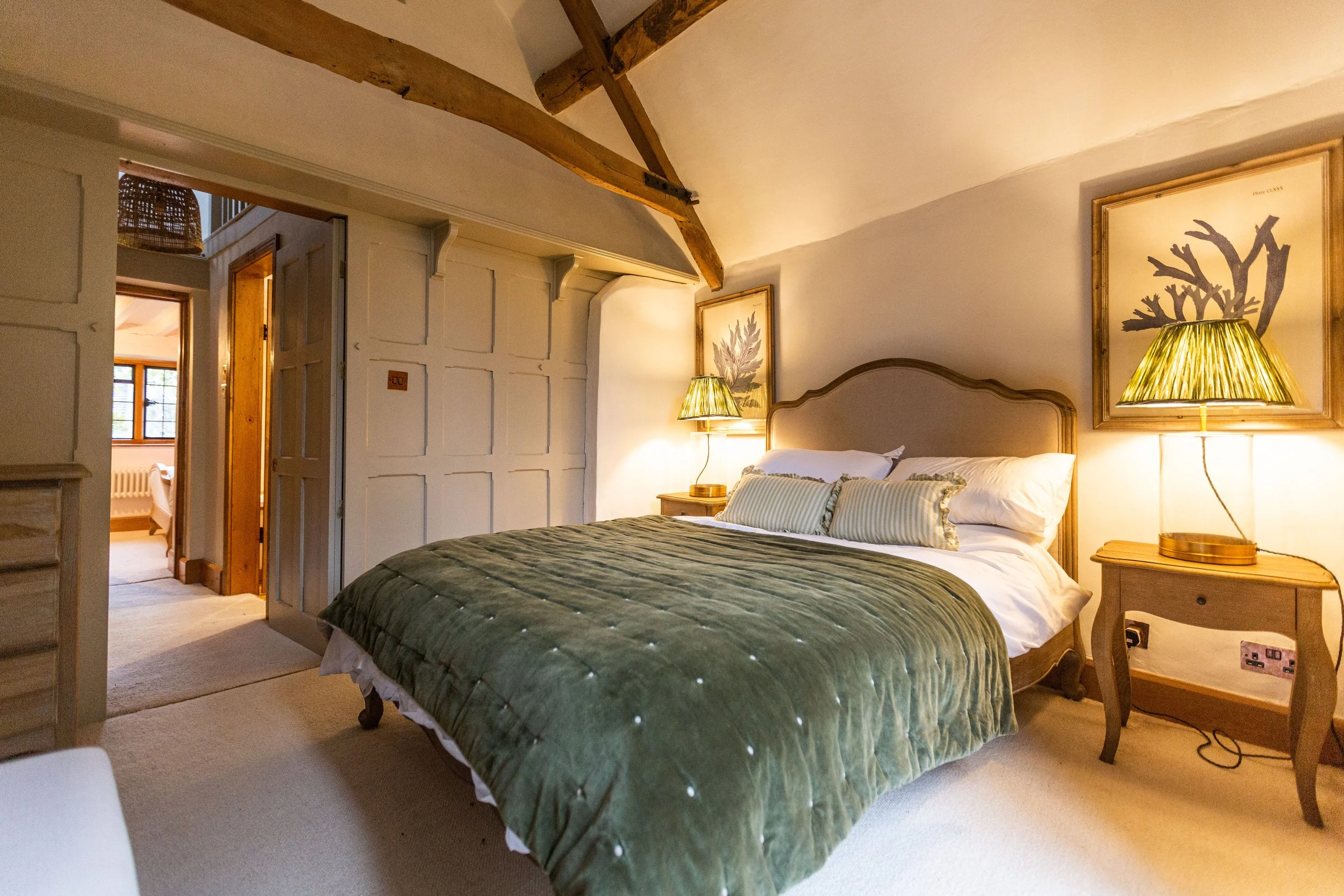 Cozy bedroom with a large bed, green velvet cover, striped pillows, bedside tables with lamps, wall art, wooden beam ceiling, and an open doorway showing another room with a bed and window.