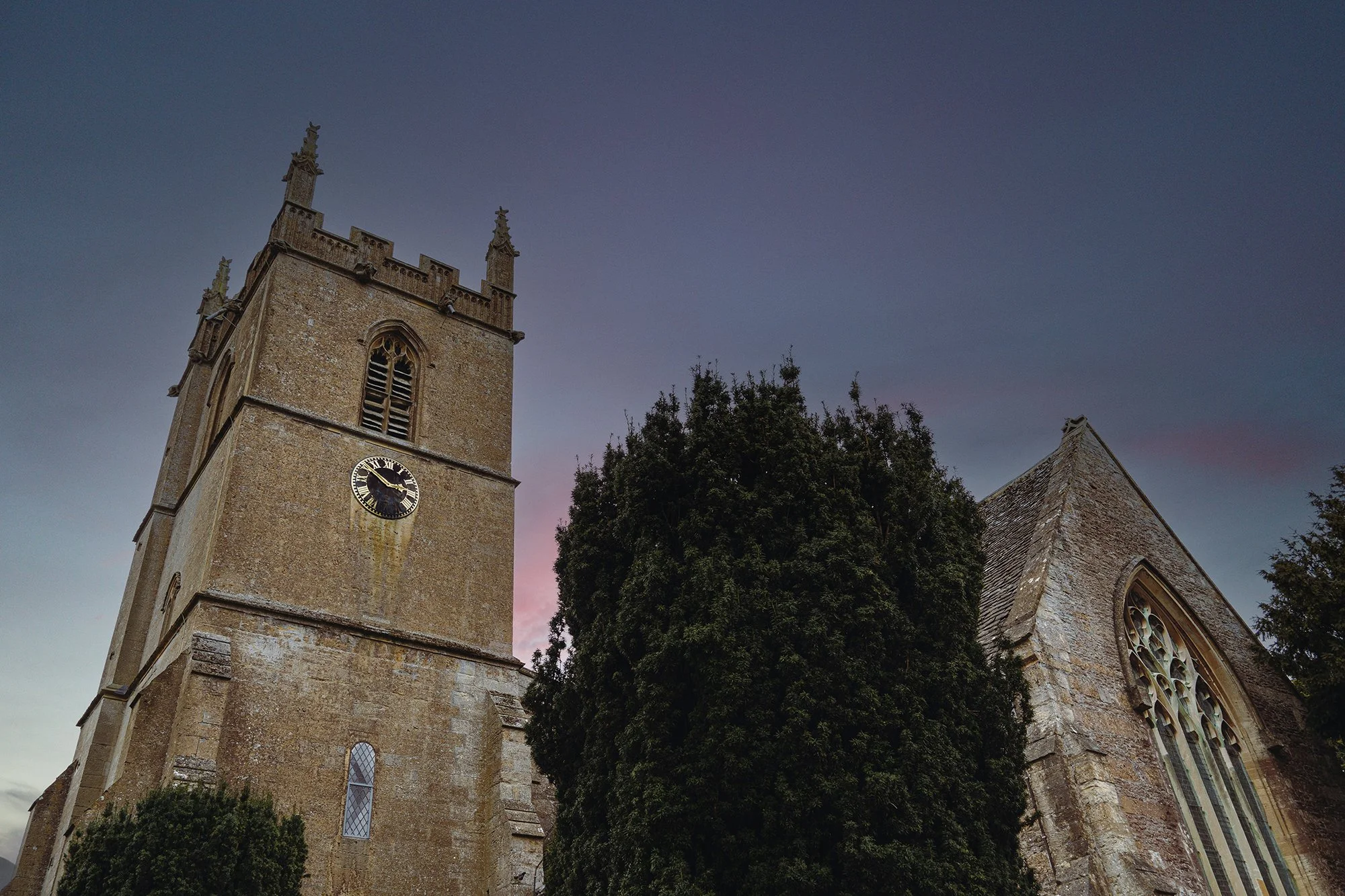 An old church with a tall clock tower and Gothic-style windows, surrounded by trees, during twilight.