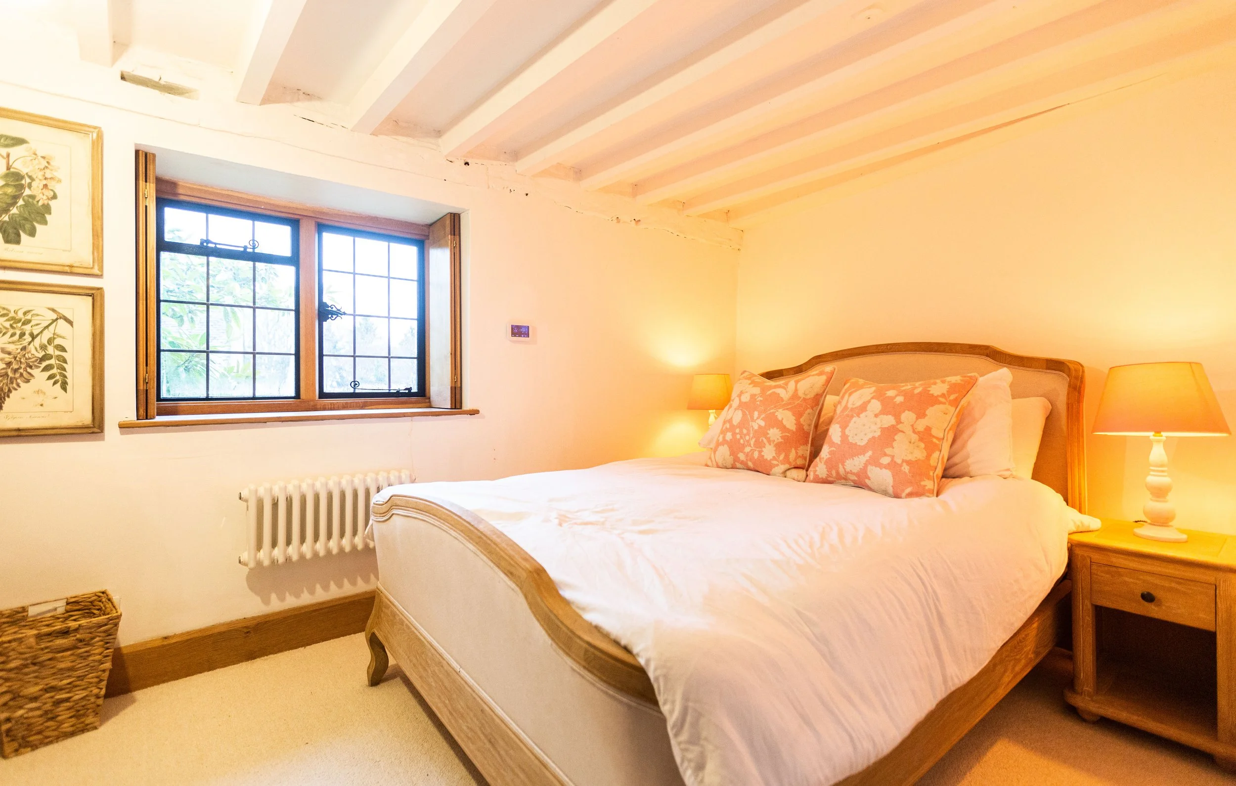 Cozy bedroom with a wooden bed frame, white bedding, pink floral pillows, bedside table with a lamp, and a window with wooden trim and leaded glass panes.