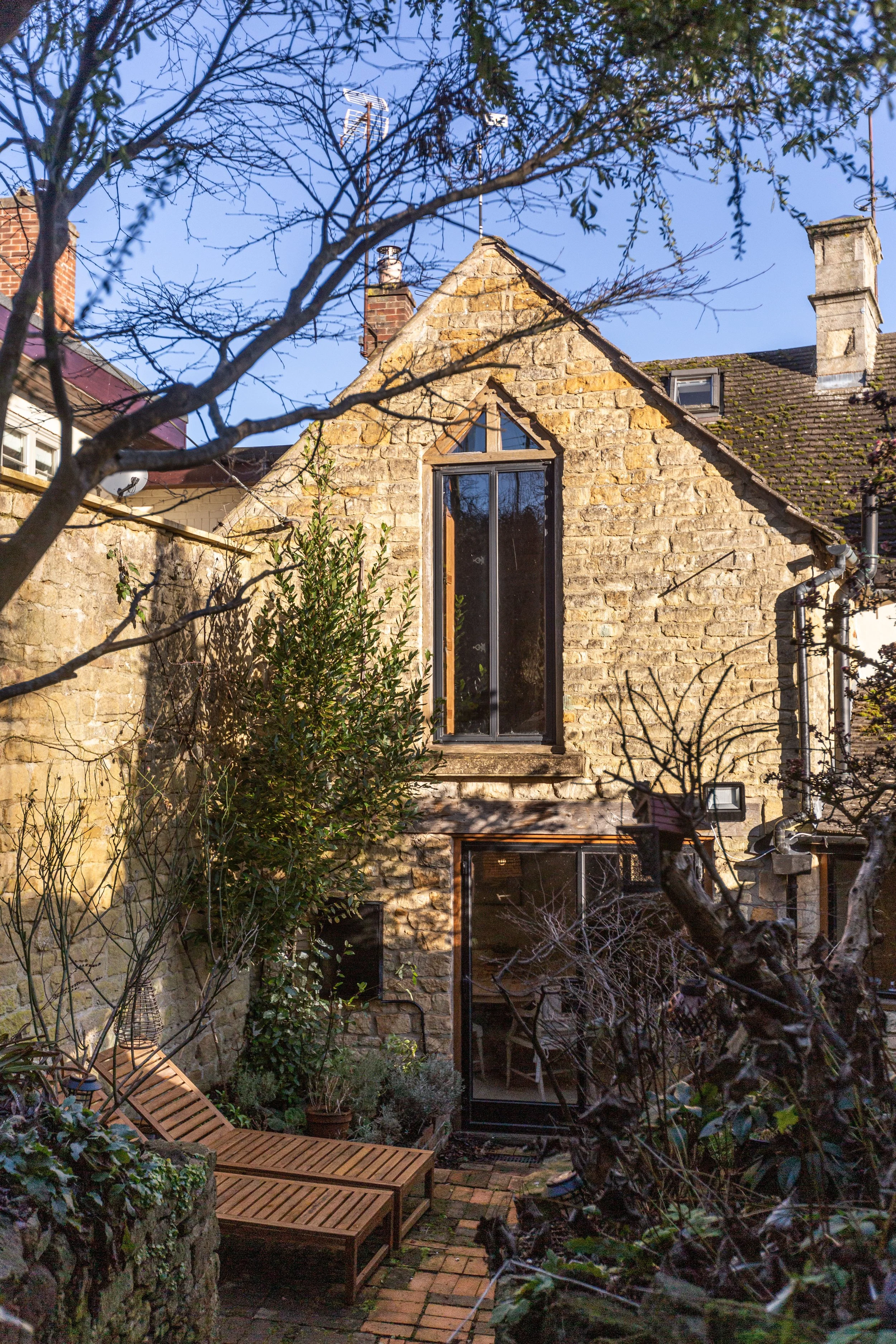 Backyard with stone house, large window, brick patio, wooden lounge chair, trees, and bushes.