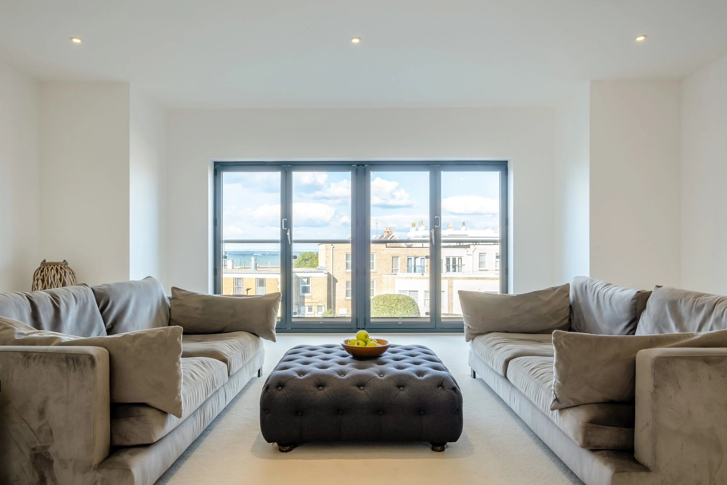 Living room with two beige sofas facing each other, a gray tufted ottoman in the center with a bowl of green apples on top, large window with a view of buildings and sky.