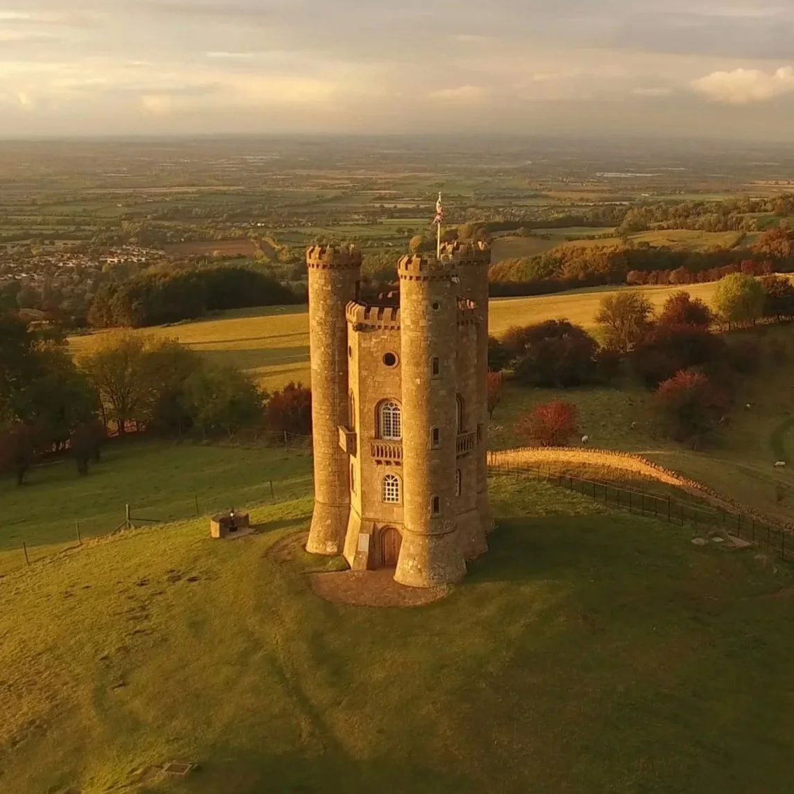 An aerial view of a medieval castle with tall cylindrical towers, situated on a grassy hill overlooking an expansive rural landscape with fields and trees in the distance during sunset.