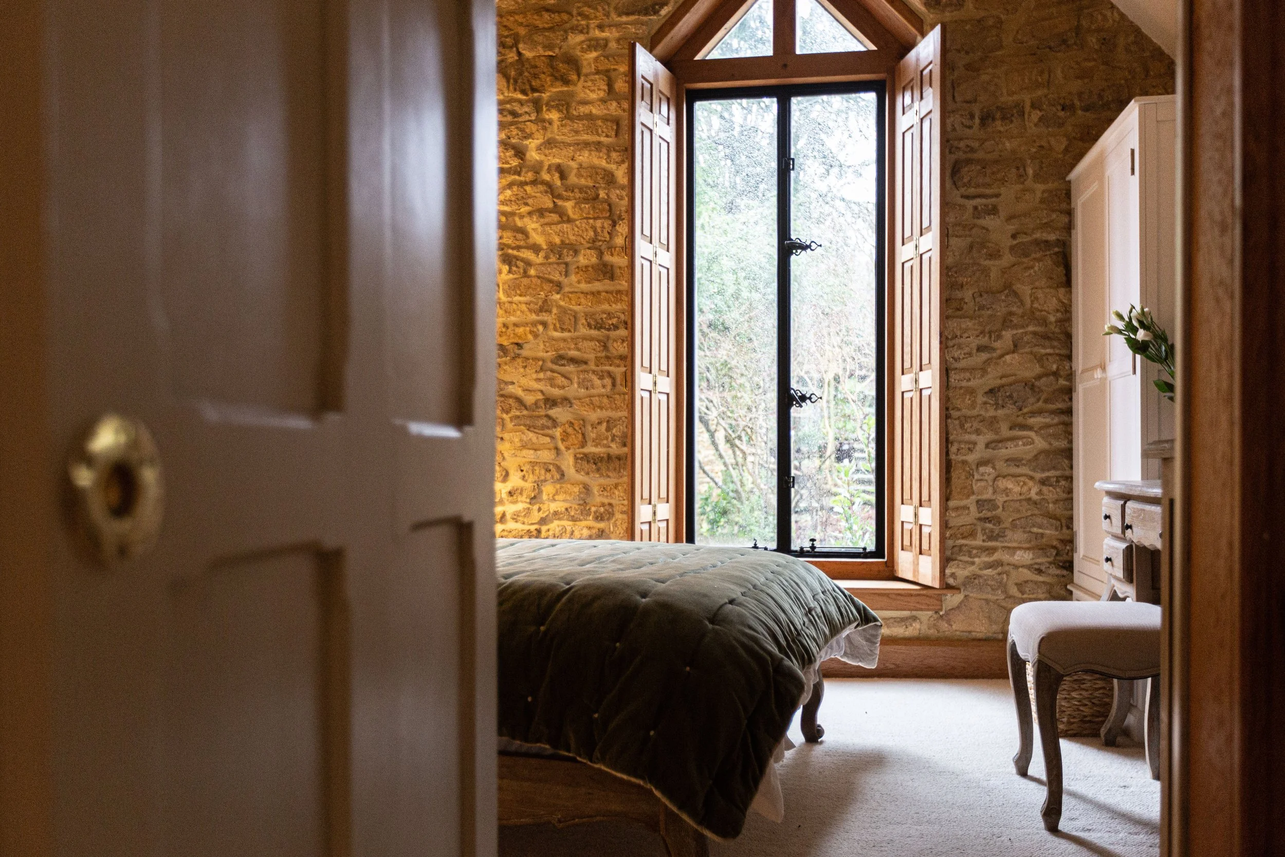 View of a bedroom with a large window with wooden shutters, exposing a garden outside. The room has stone walls, a bed with a dark comforter, a white cabinet, and a small table with a plant. An open door frames the scene.