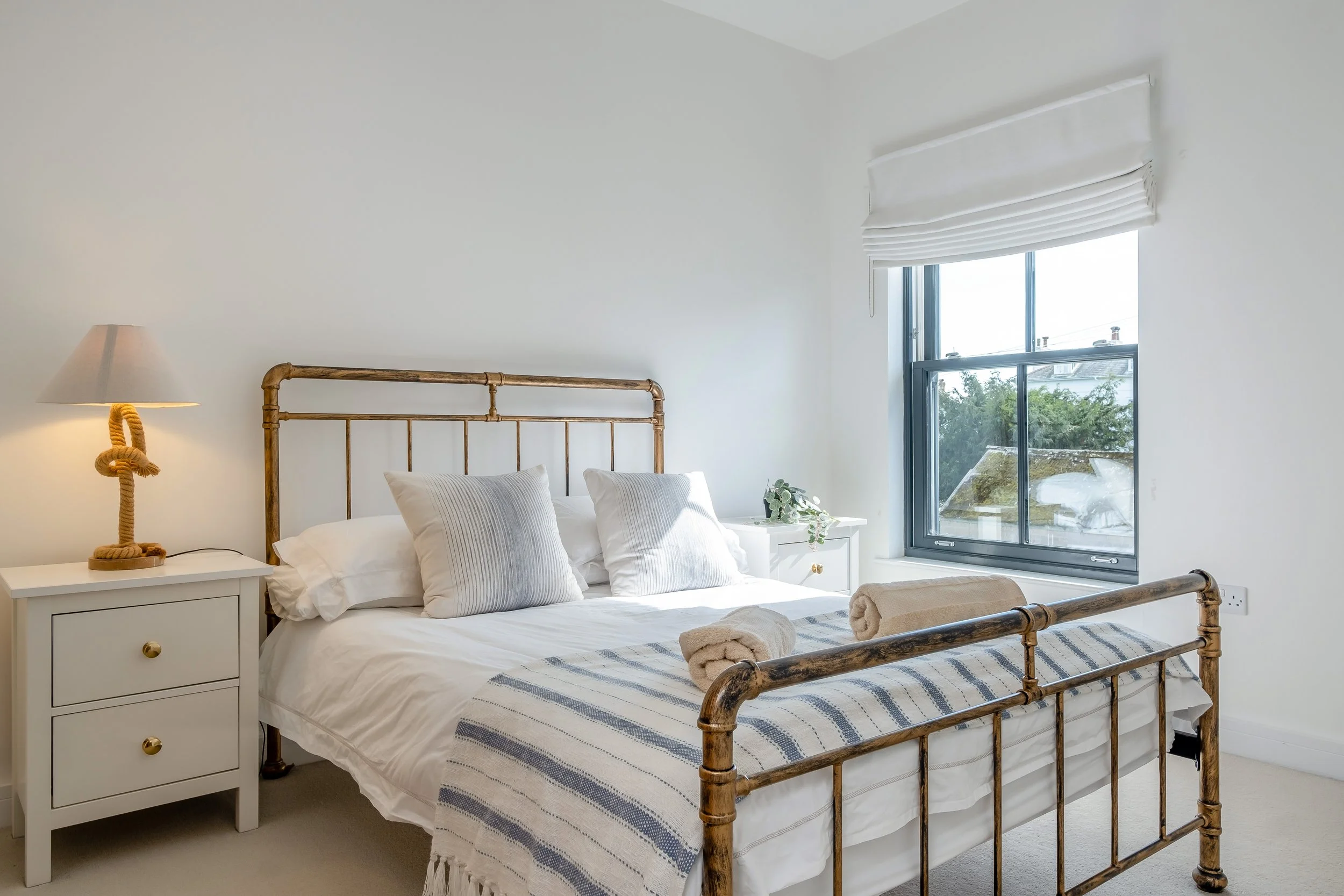 A minimalist bedroom with a white bed, wooden headboard, and two white nightstands. One nightstand has a beige lamp and the other has a small plant. The bed has white sheets, striped pillows, and folded towels. There is a window with a white blind.