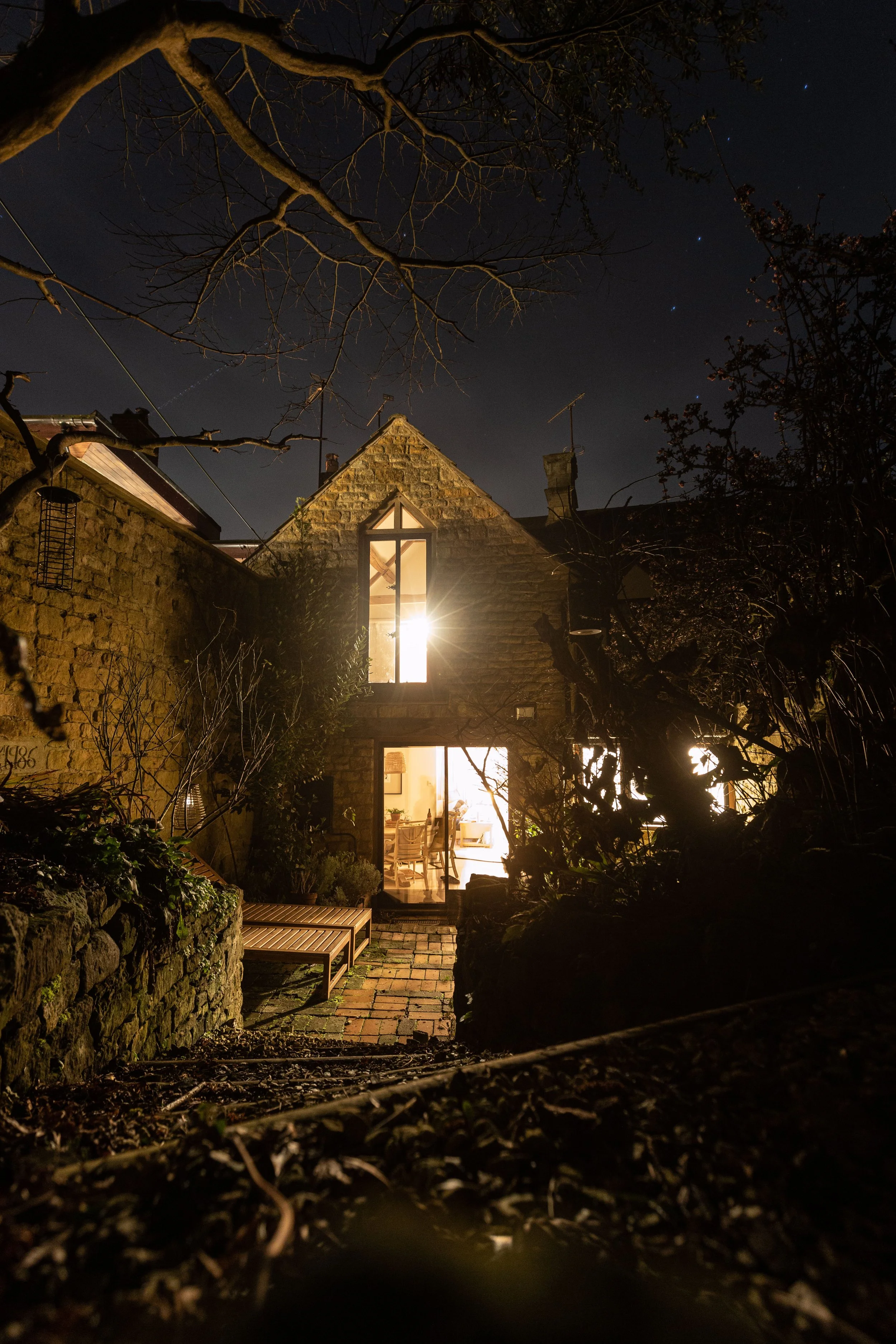 Nighttime view of a house with an illuminated interior, visible through large glass doors and windows. The house is surrounded by trees and a stone pathway leading to the entrance.