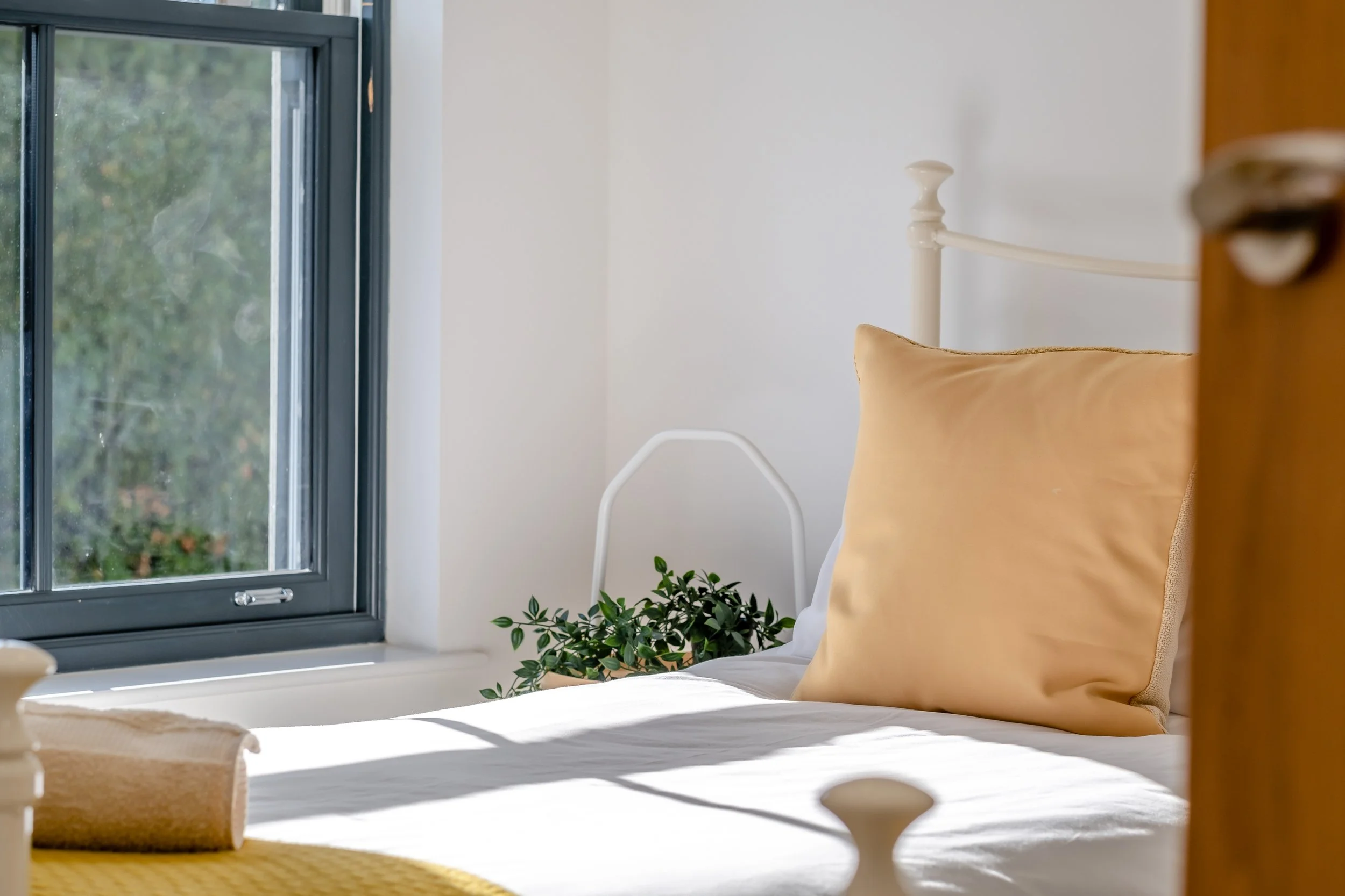 A bedroom with a white bed frame, beige pillow, and white bedding, positioned near a window with sunlight casting shadows, and a small green plant on the windowsill.