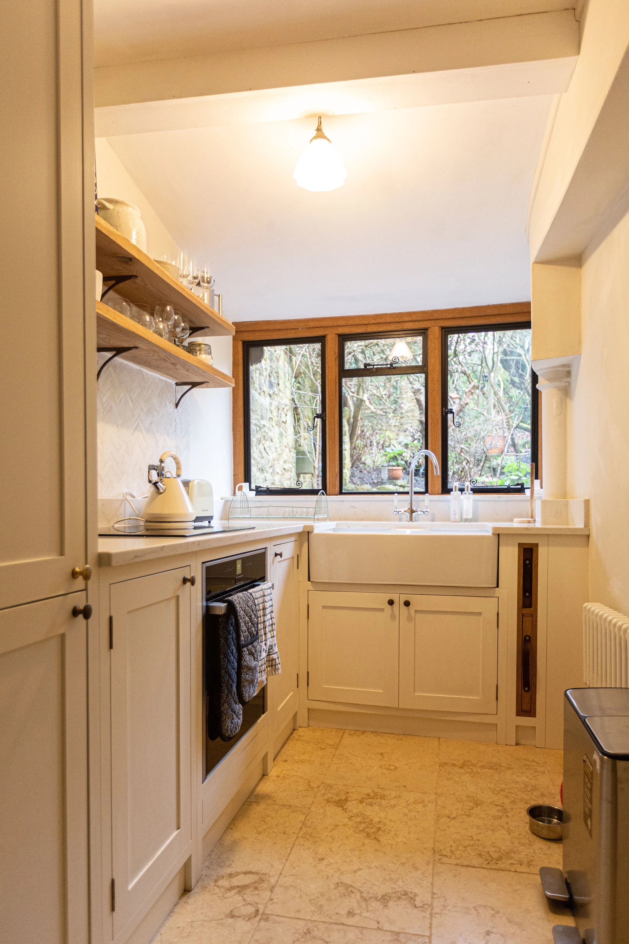 A cozy kitchen with cream-colored cabinets, a white farmhouse sink, and open wooden shelves displaying glassware. A window over the sink shows a view of trees outside.