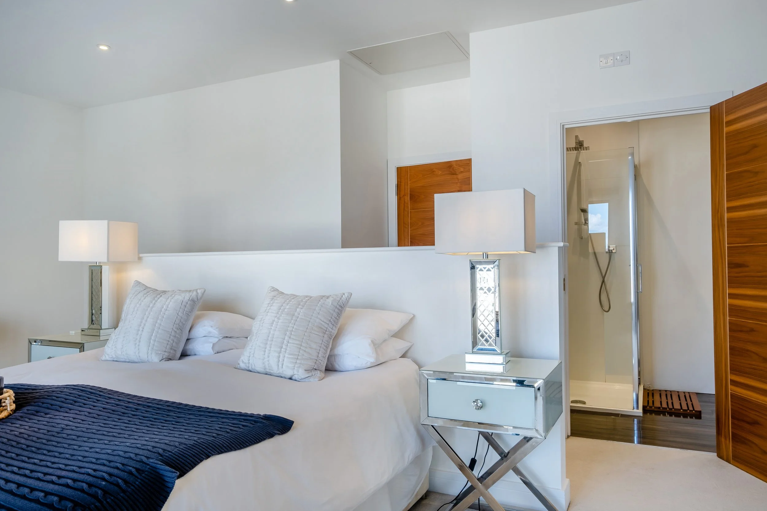 A neatly made bed with white linens and pillows in a modern bedroom. Two mirrored bedside tables with lamps are on each side of the bed. An open bathroom door reveals a shower with a glass door and a handheld showerhead.