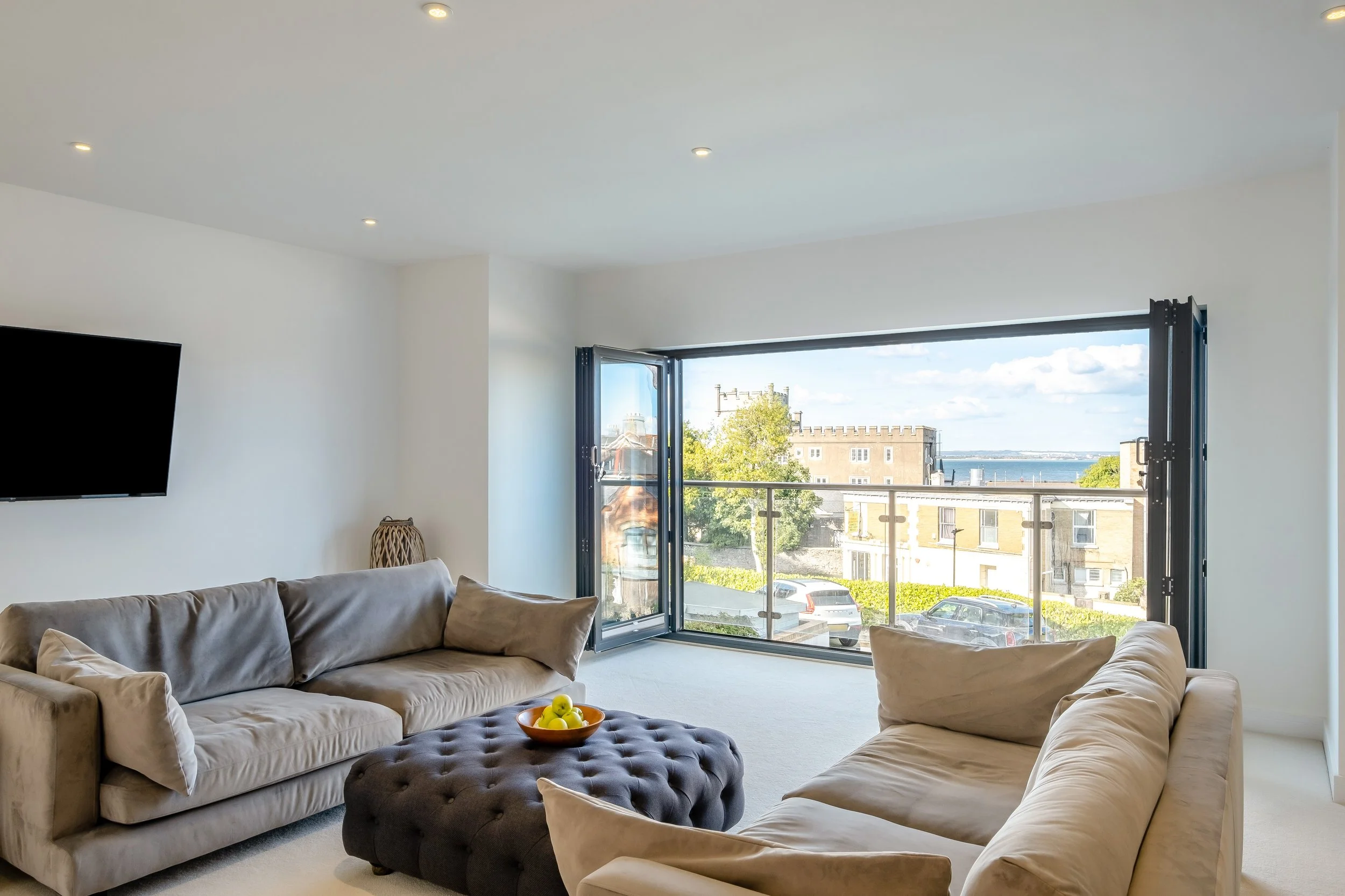 Living room with beige sofas, a tufted ottoman with a yellow fruit bowl, large open window with a view of buildings and water, television on the wall, and minimal decor.