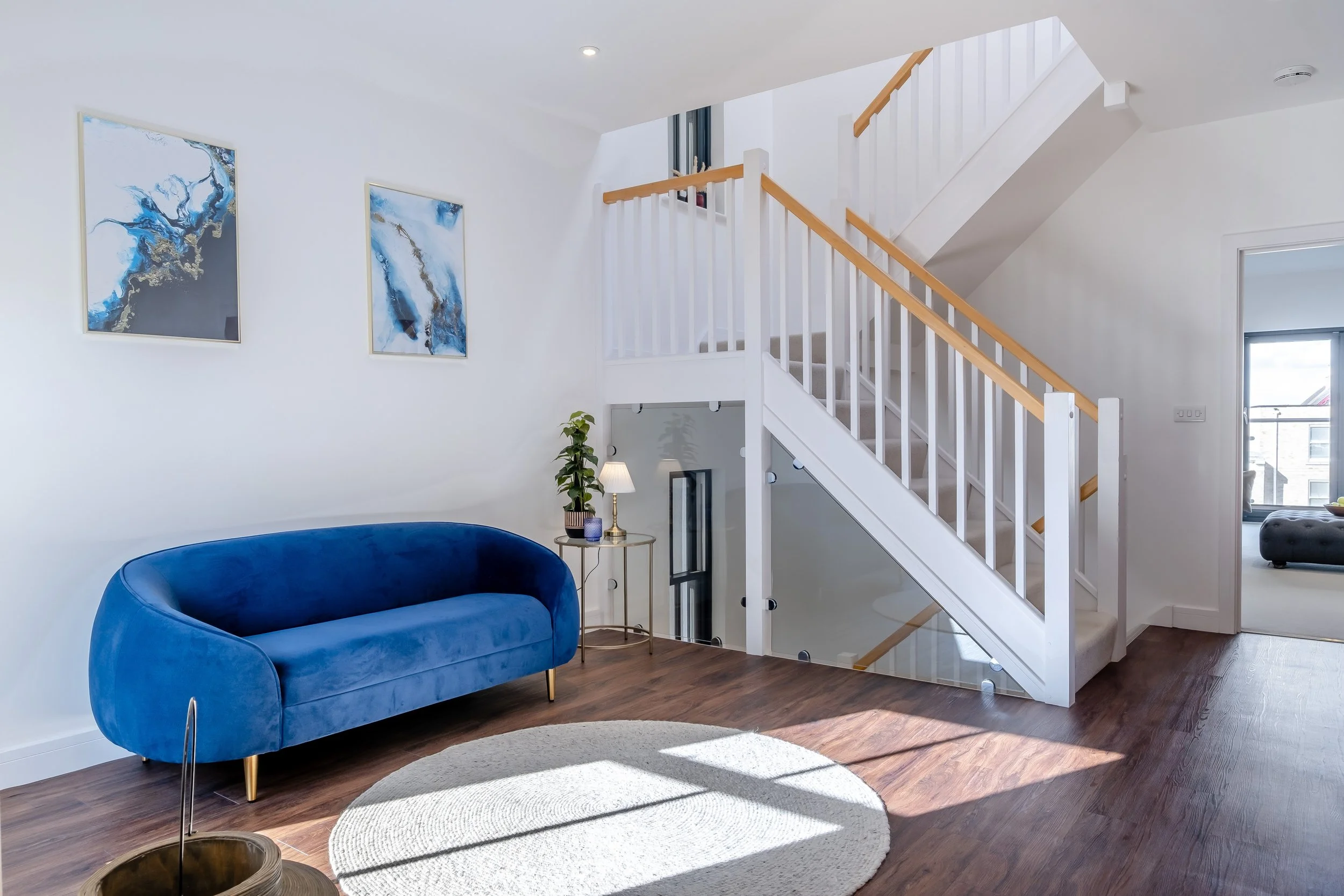 Living room with blue sofa, wall art, small side table with lamp and plant, staircase with glass railing, and sunlight on wooden floor.
