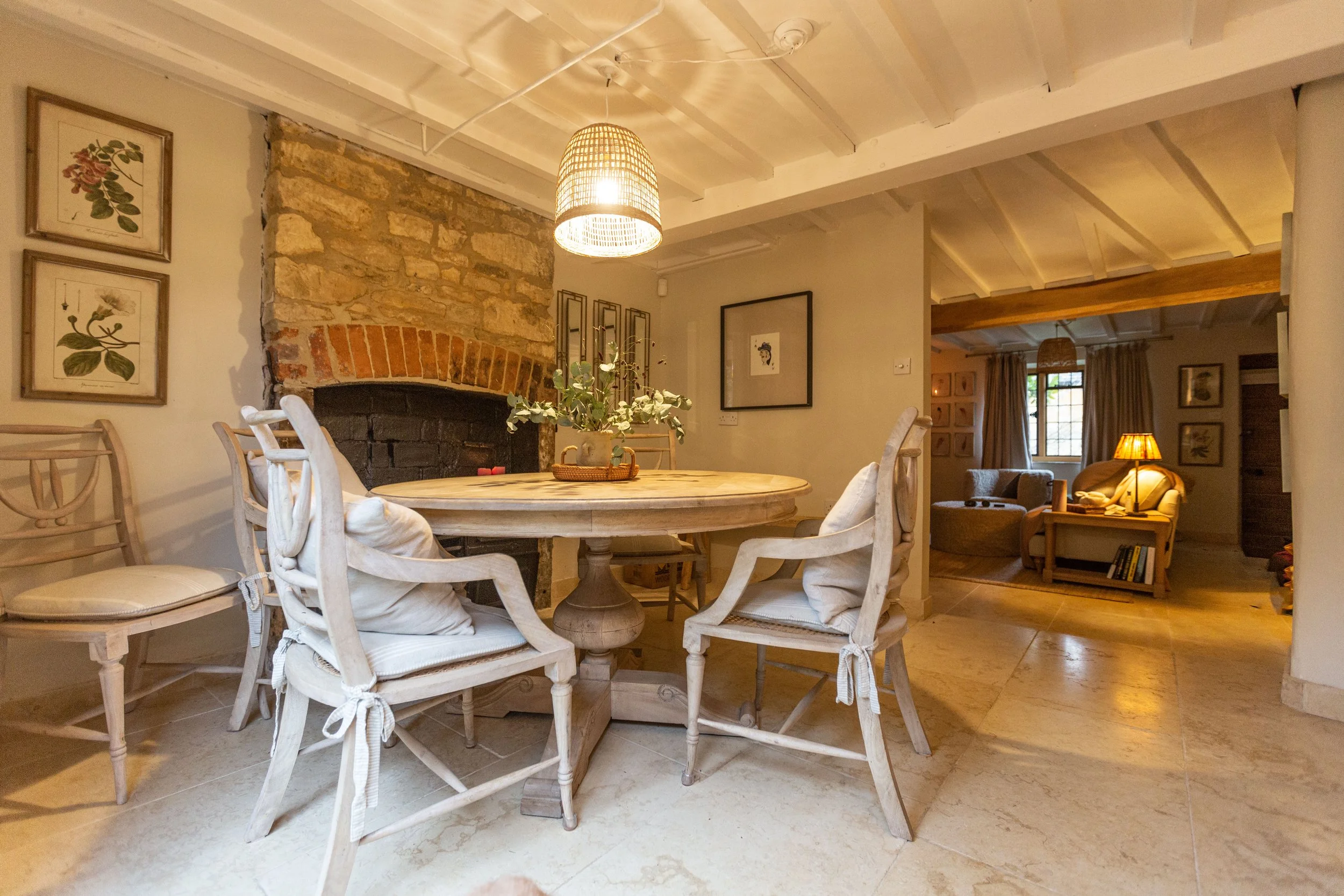 A cozy dining area with a round wooden table surrounded by four white chairs with cushions. There is a plant in the center of the table, and a rustic stone and brick fireplace in the background. The room features wooden beams, framed botanical prints on the walls, and warm ambient lighting.