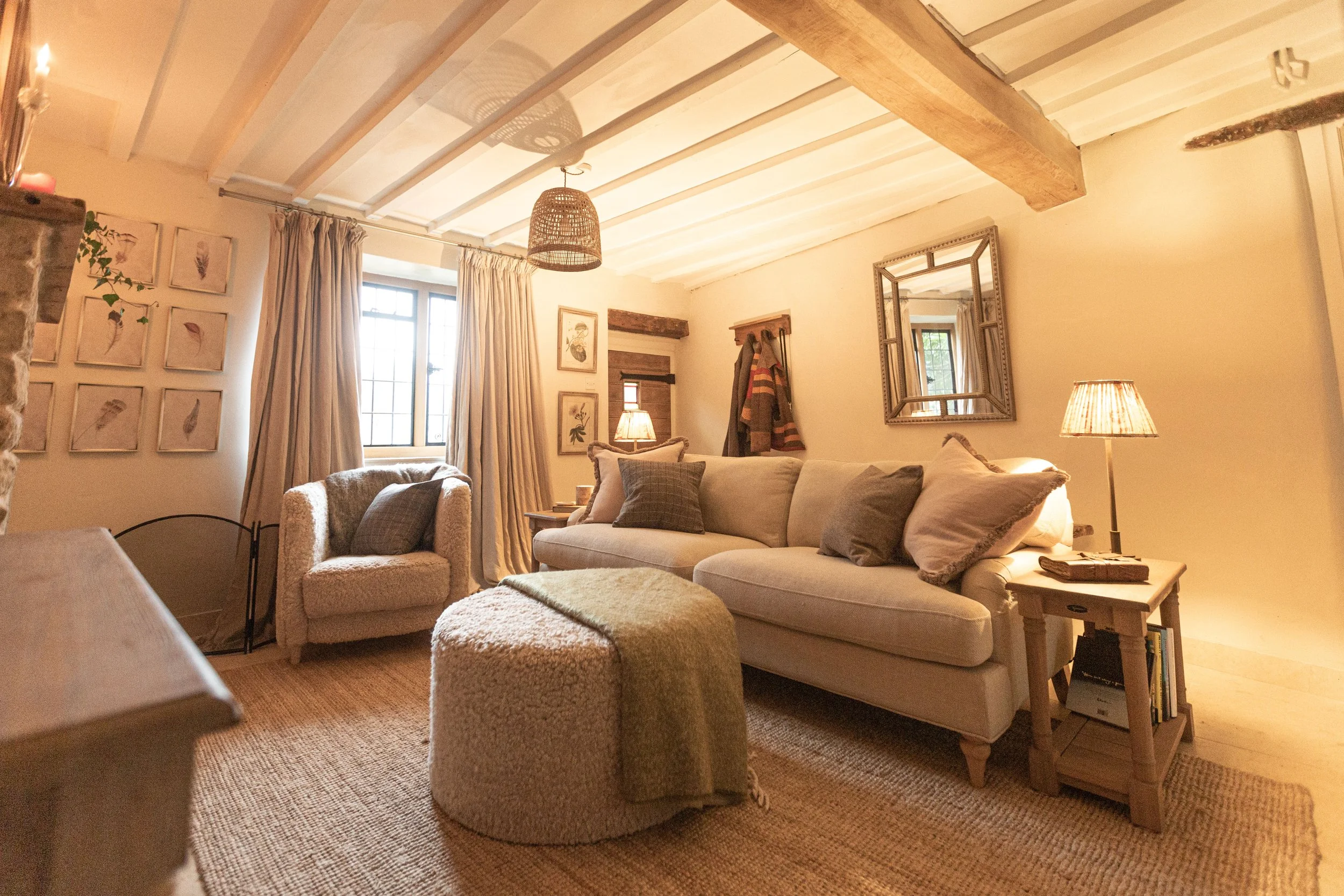 Cozy living room with beige sofa and armchair, wooden side table with books, framed wall art, window with beige curtains, patterned ottoman, and warm lighting.