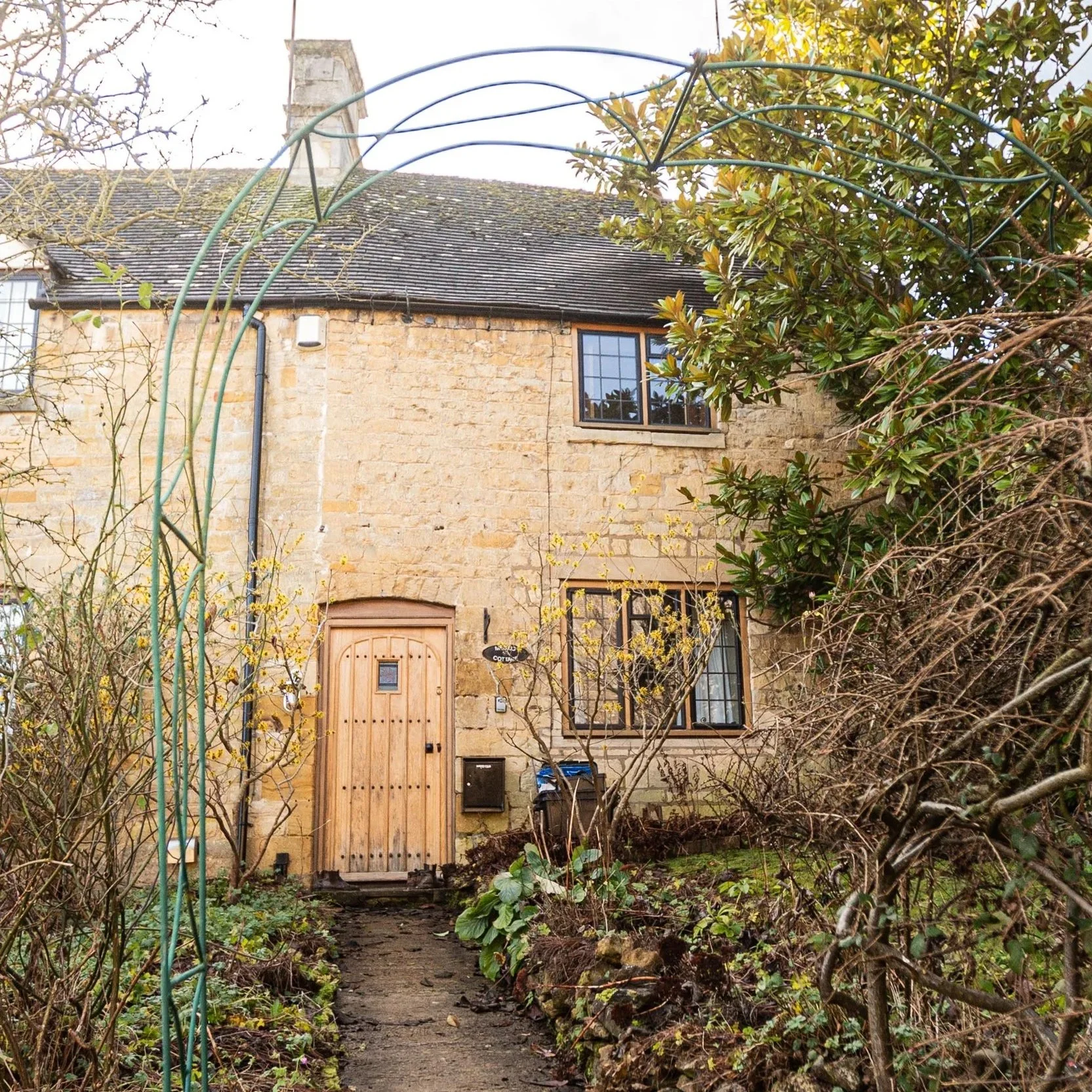 A stone house with a wooden front door, two windows with dark frames, a small garden with plants and trees, and a metal garden archway in the foreground.
