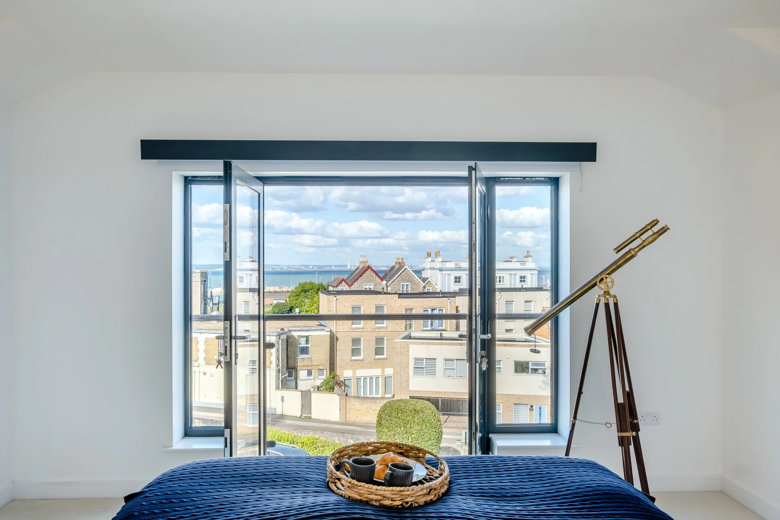 Bedroom with large open window, view of cityscape, ocean in the distance, telescope on stand, bed with dark blue cover, tray with teacups on bed, bright and airy room