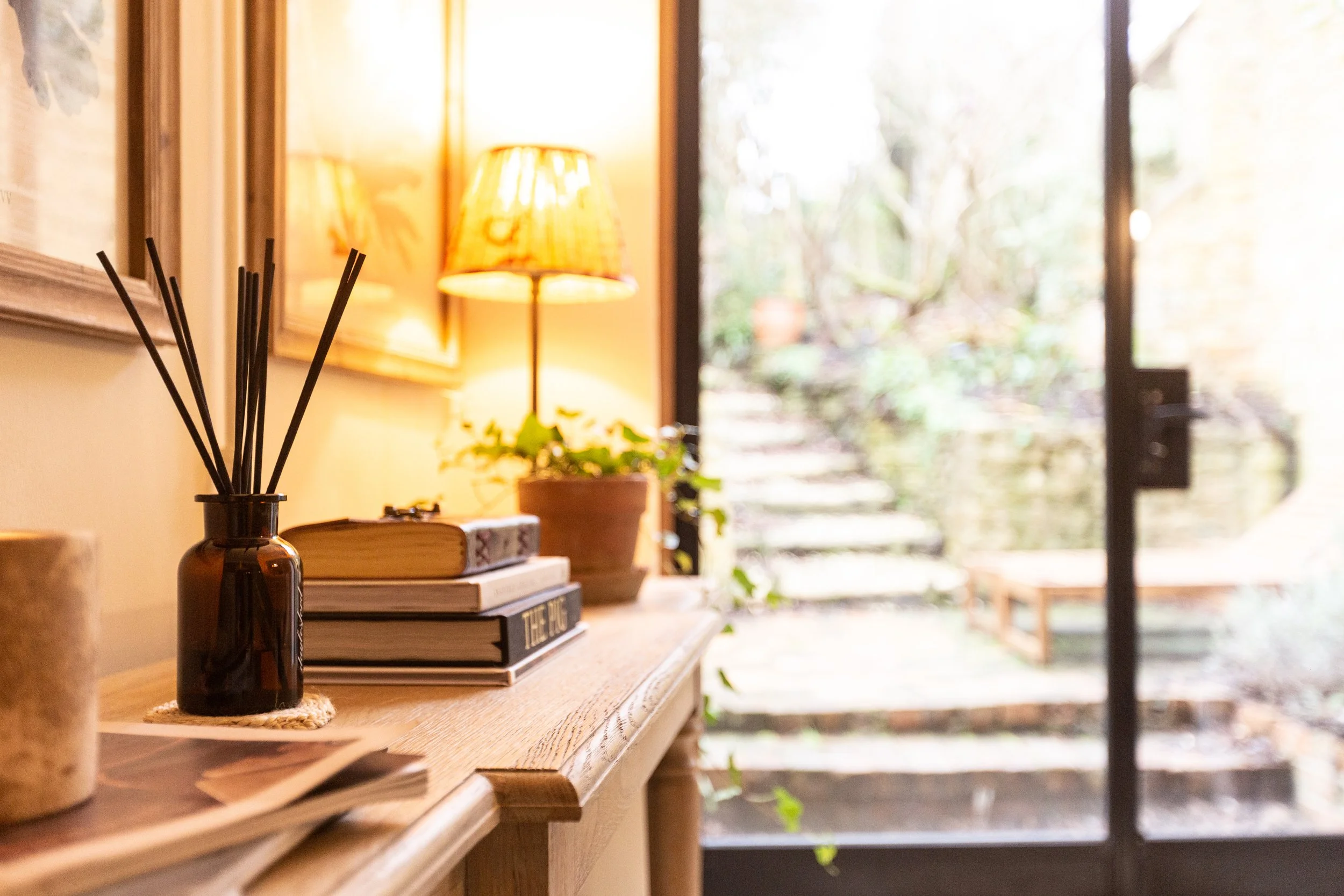 Interior scene with a wooden table holding a black reed diffuser, books, and a potted plant, with a warm table lamp and a garden visible through a glass door in the background.