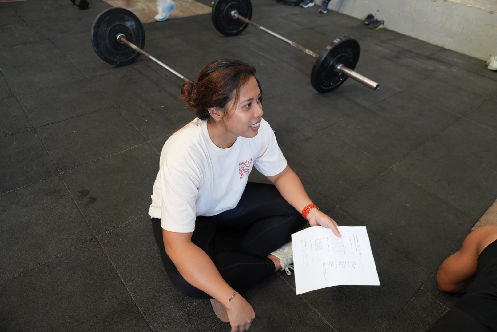 A woman sitting on the gym floor holding a paper, with barbell weights in the background.