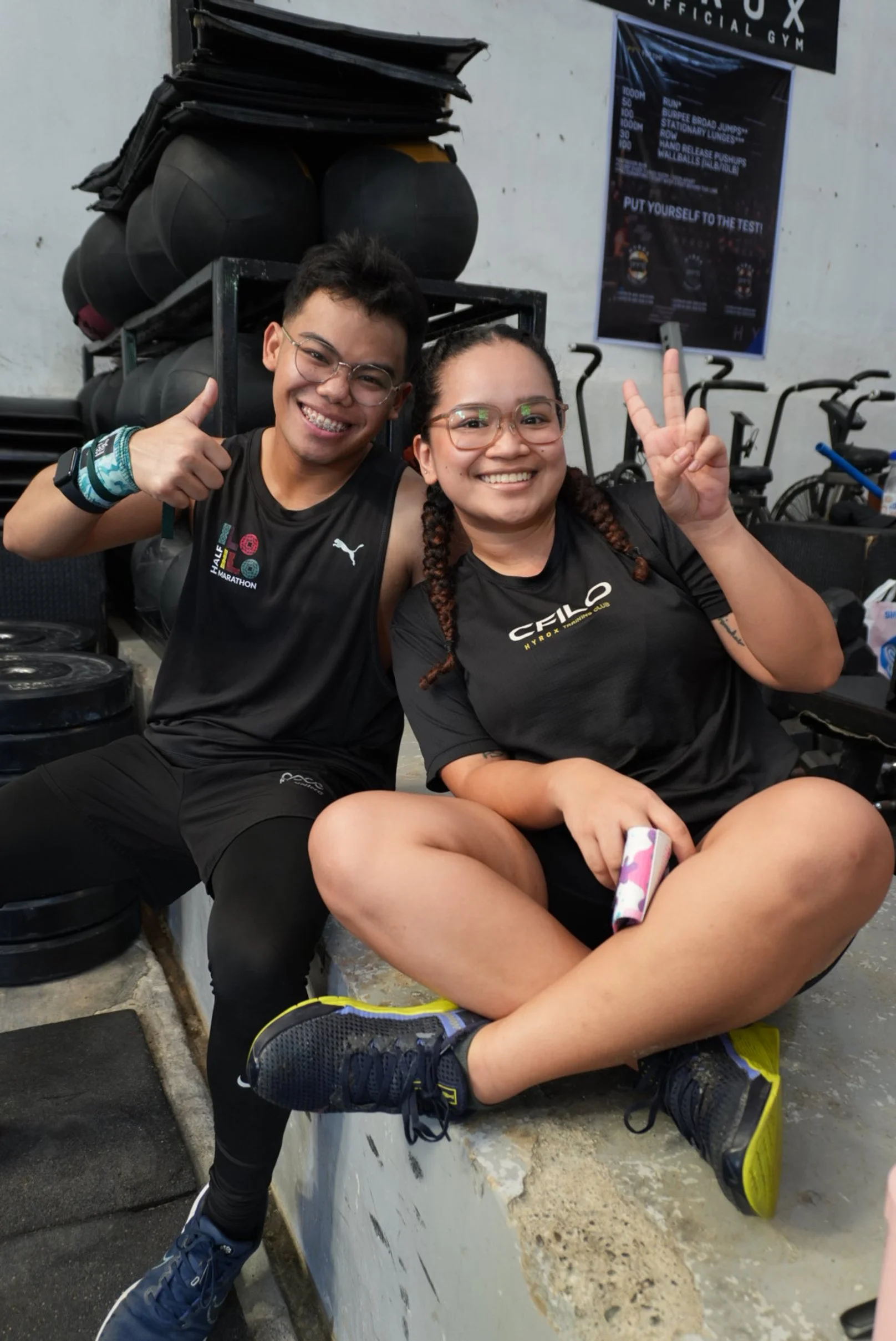 Two smiling people at a gym, one giving a thumbs-up and the other making a peace sign, sitting on a gym bench with workout equipment behind them.