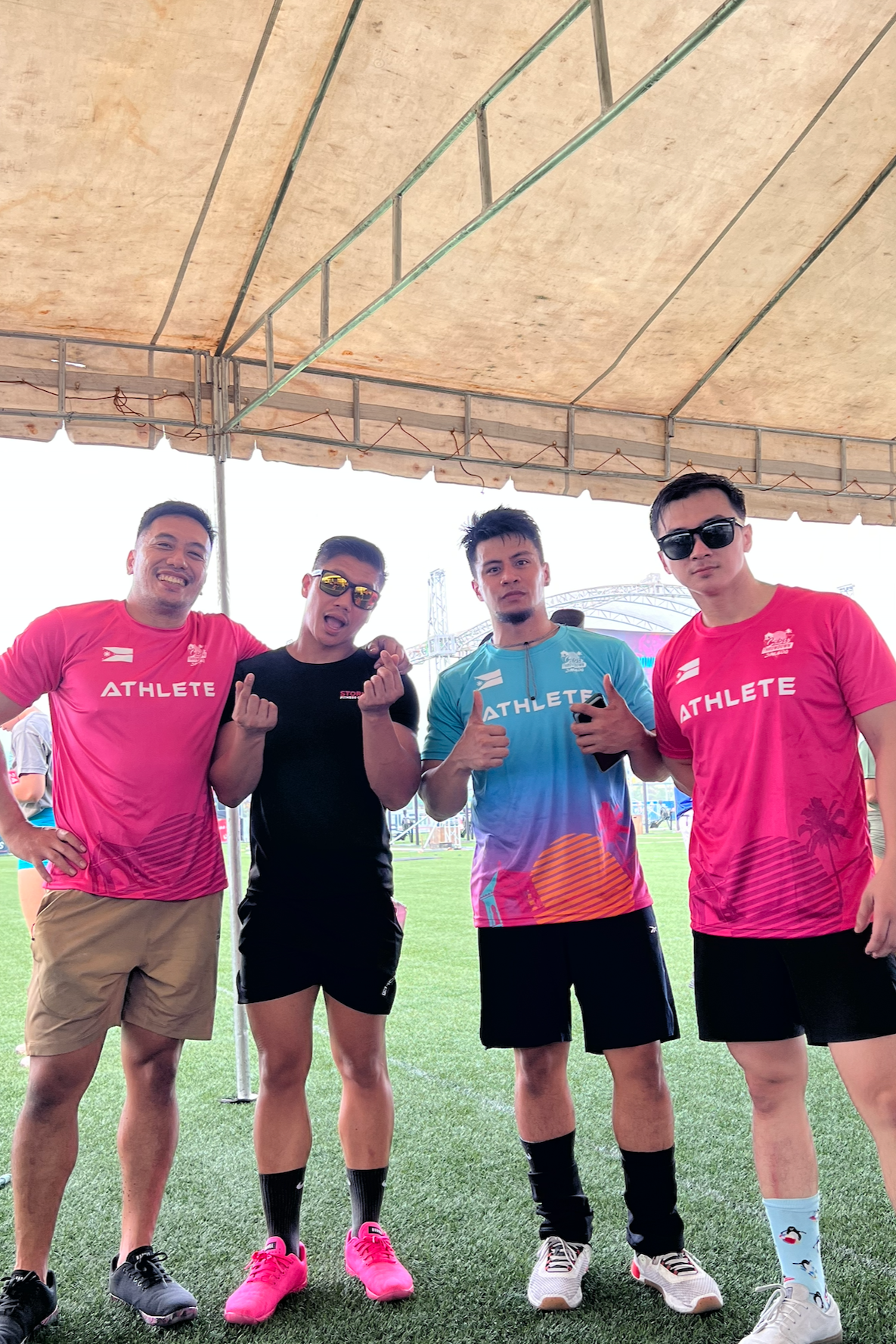 Four men standing under a tent at a sports field, wearing athletic shirts with 'ATHLETE' printed on them, showing cheerful and playful gestures.