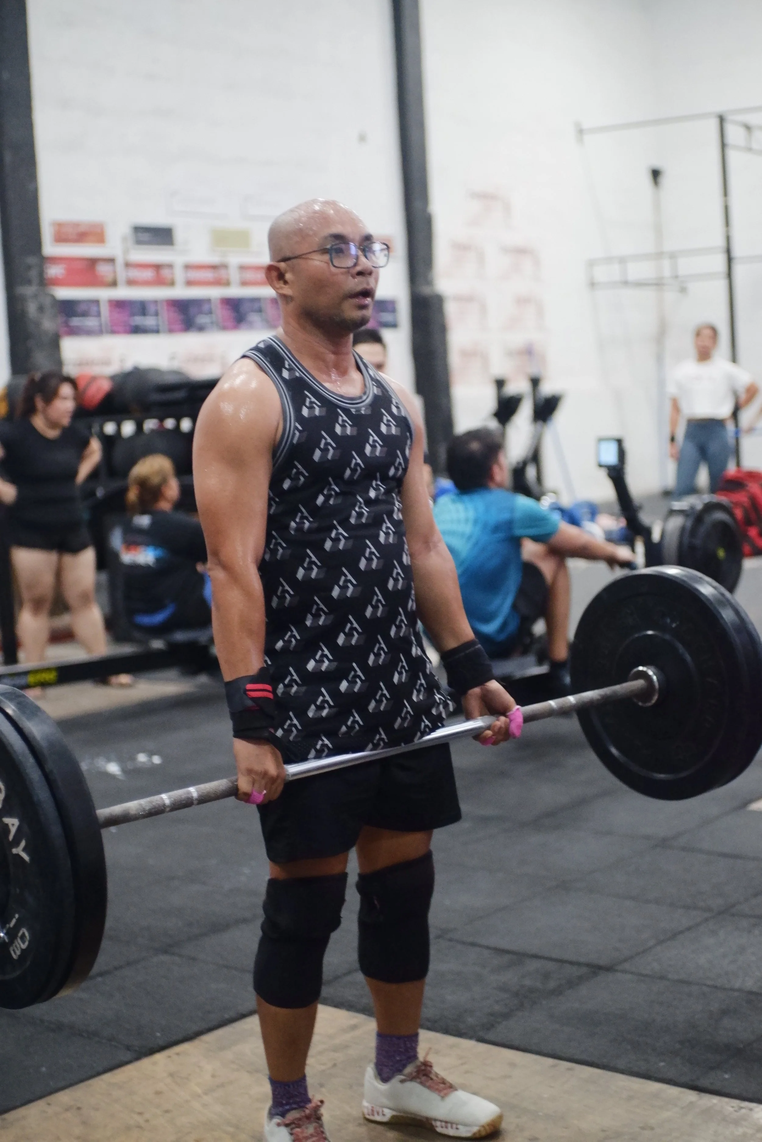 A man lifting a barbell at a gym or CrossFit event, surrounded by people, with weights on each side of the barbell, wearing workout attire and knee sleeves.