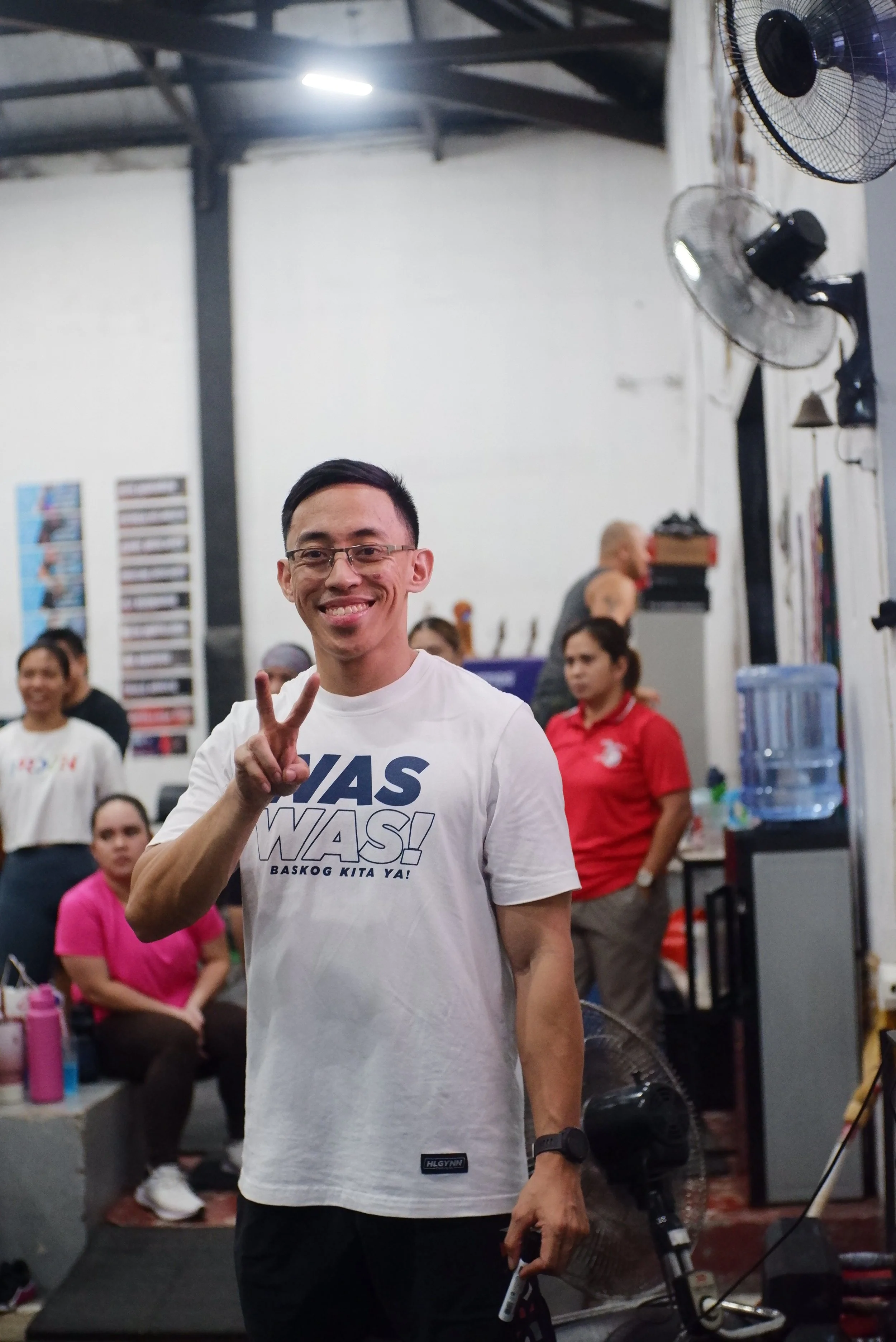 A young man smiling and making a peace sign in an indoor space, with several people and objects in the background, including wall fans and a water cooler.