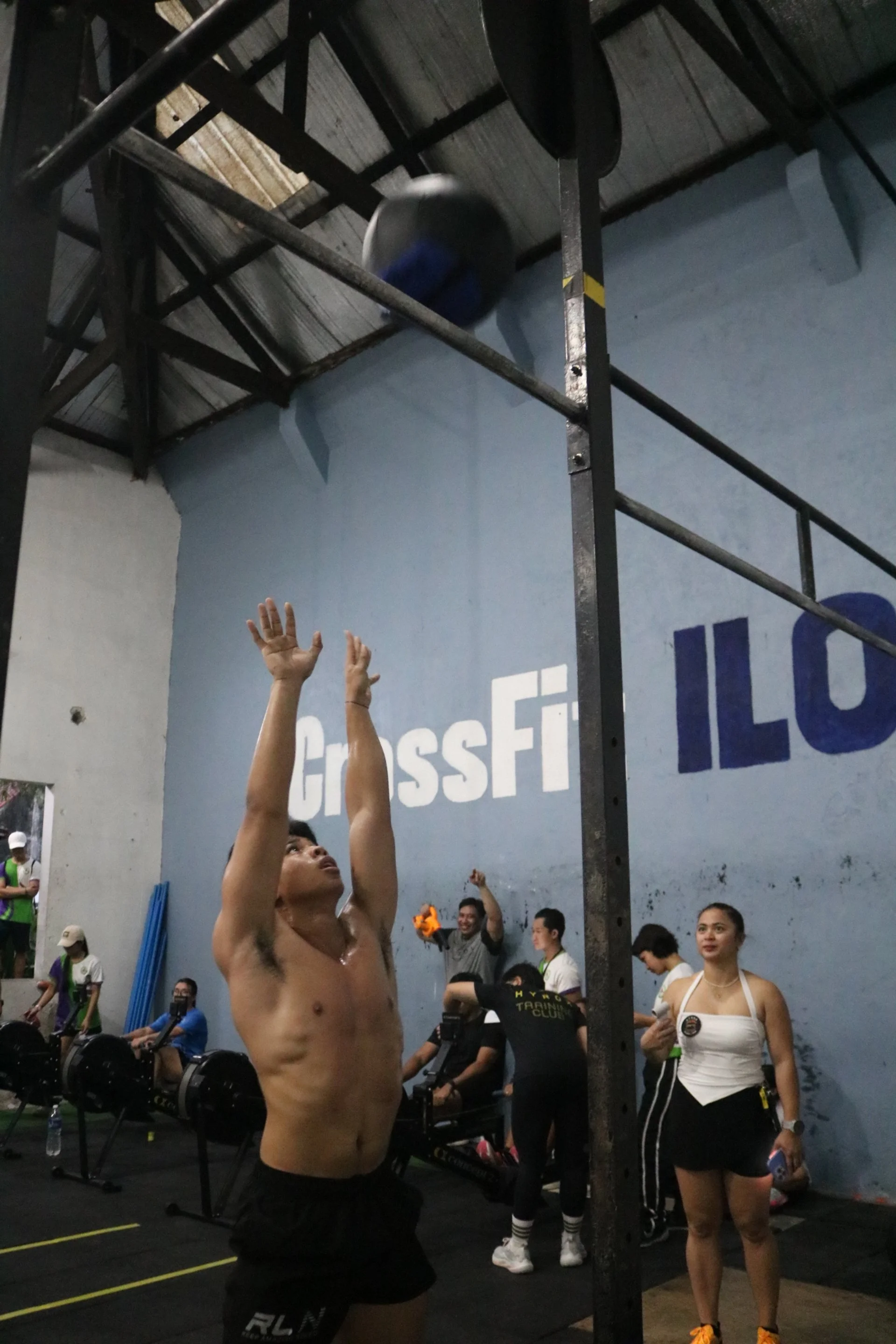 A male athlete with a muscular build and wearing black shorts is reaching up towards a gray weighted ball on a vault. The background shows a CrossFit gym with various equipment, other people, and a large blue wall with the word 'CrossFit' painted on it.