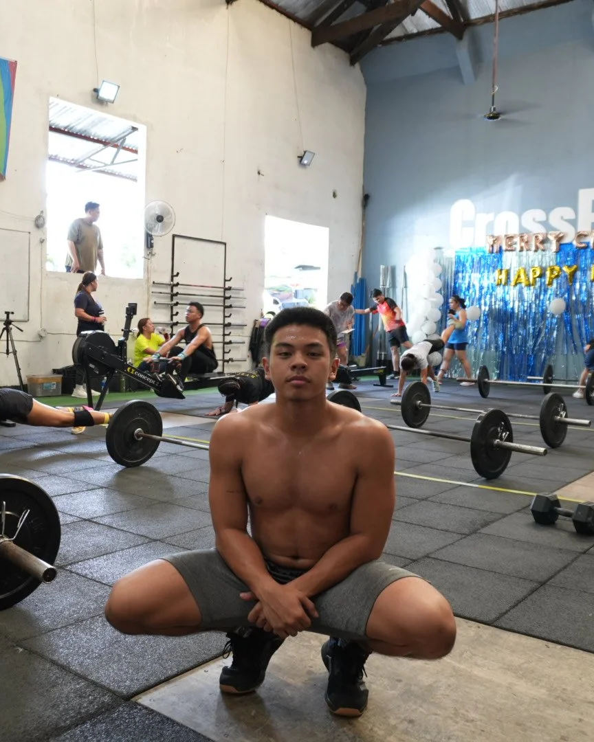 A shirtless young man crouching in the foreground of a gym with weightlifting equipment, people exercising and socializing in the background, and a decorative wall with balloons and signs.
