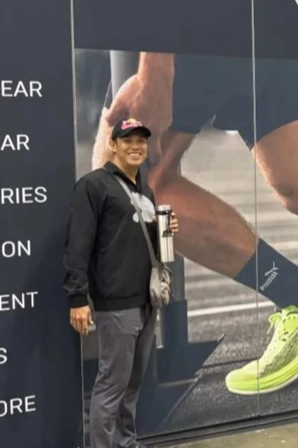 A man smiling and standing in front of a large motivational sports poster, holding a water bottle, in an indoor setting.