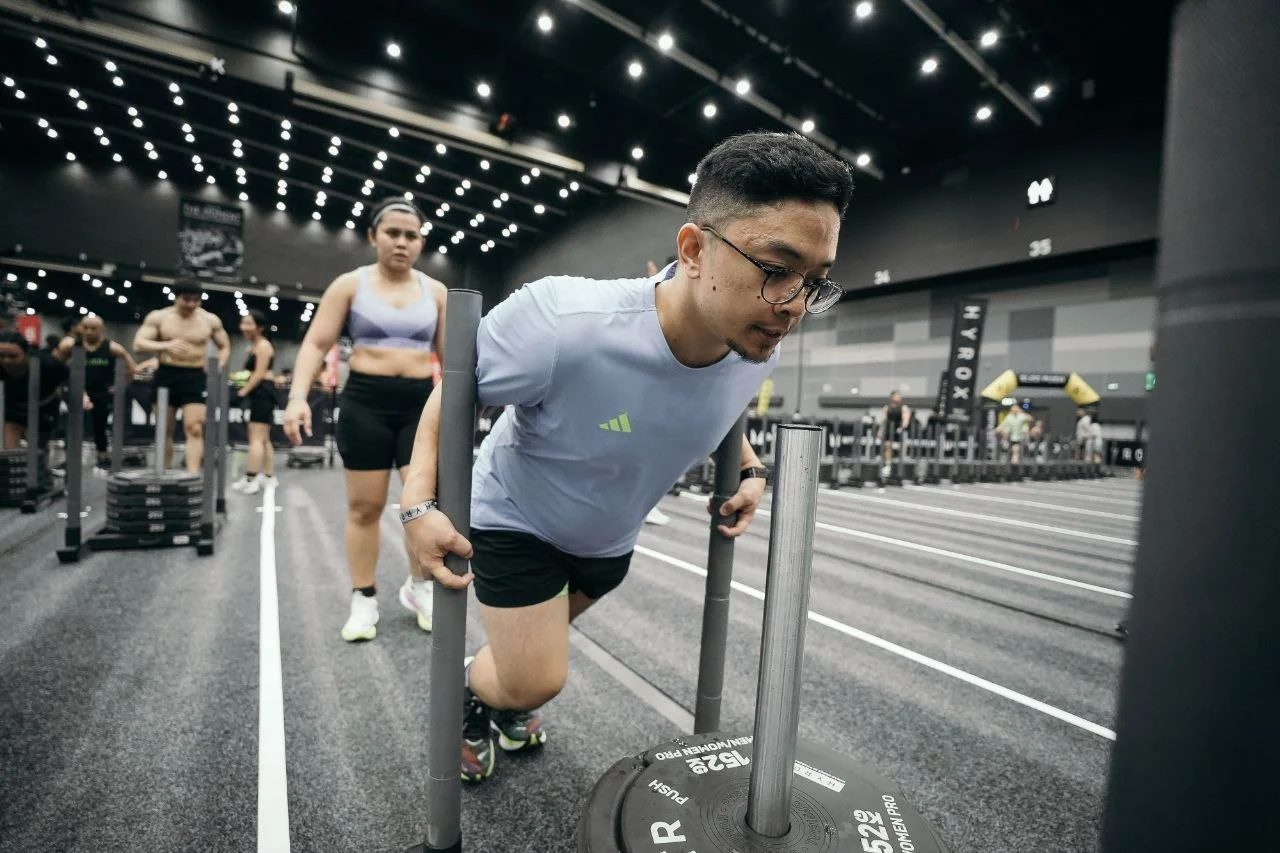 A man in a white t-shirt and black shorts pushing a weighted sled during a workout at a gym, with other people exercising in the background.