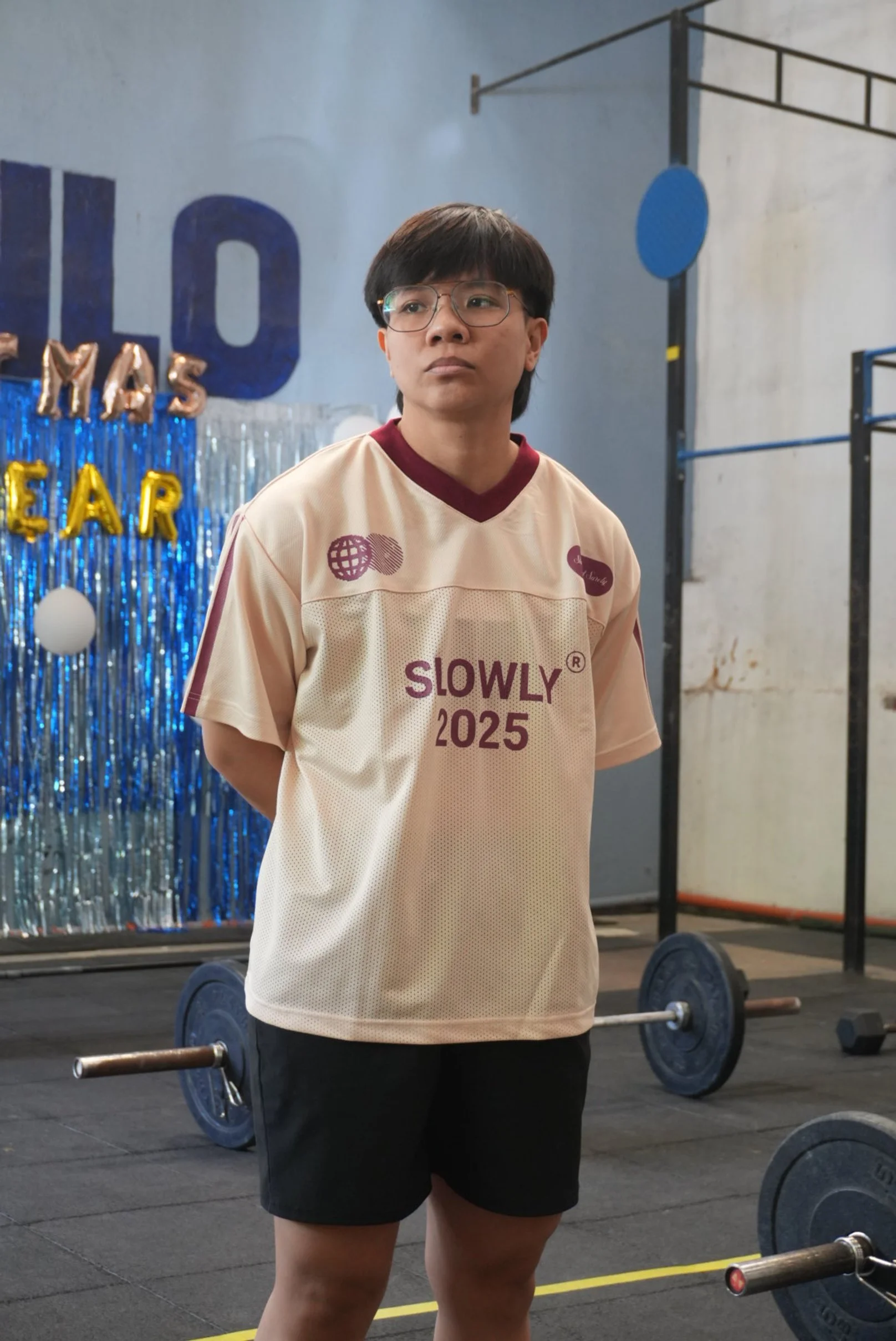 A young man wearing glasses and a beige sports shirt with 'SLOWLY 2025' printed on it, standing in a gym with weightlifting equipment in the background, during a celebration event with blue and gold balloons and metallic streamers.