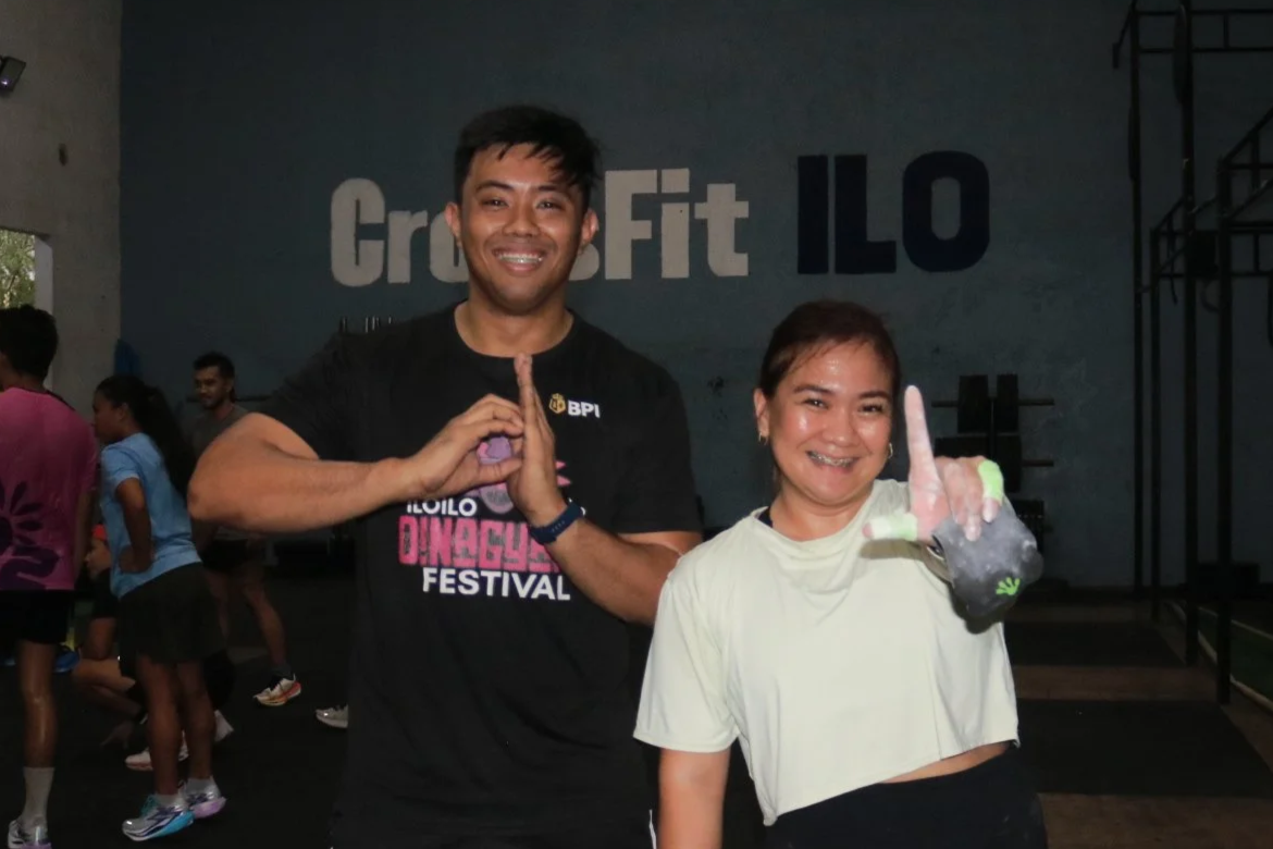 A man and woman smiling at a CrossFit event, with the man making a prayer gesture and the woman holding up her hand in a fist, both standing inside a gym with other participants in the background.