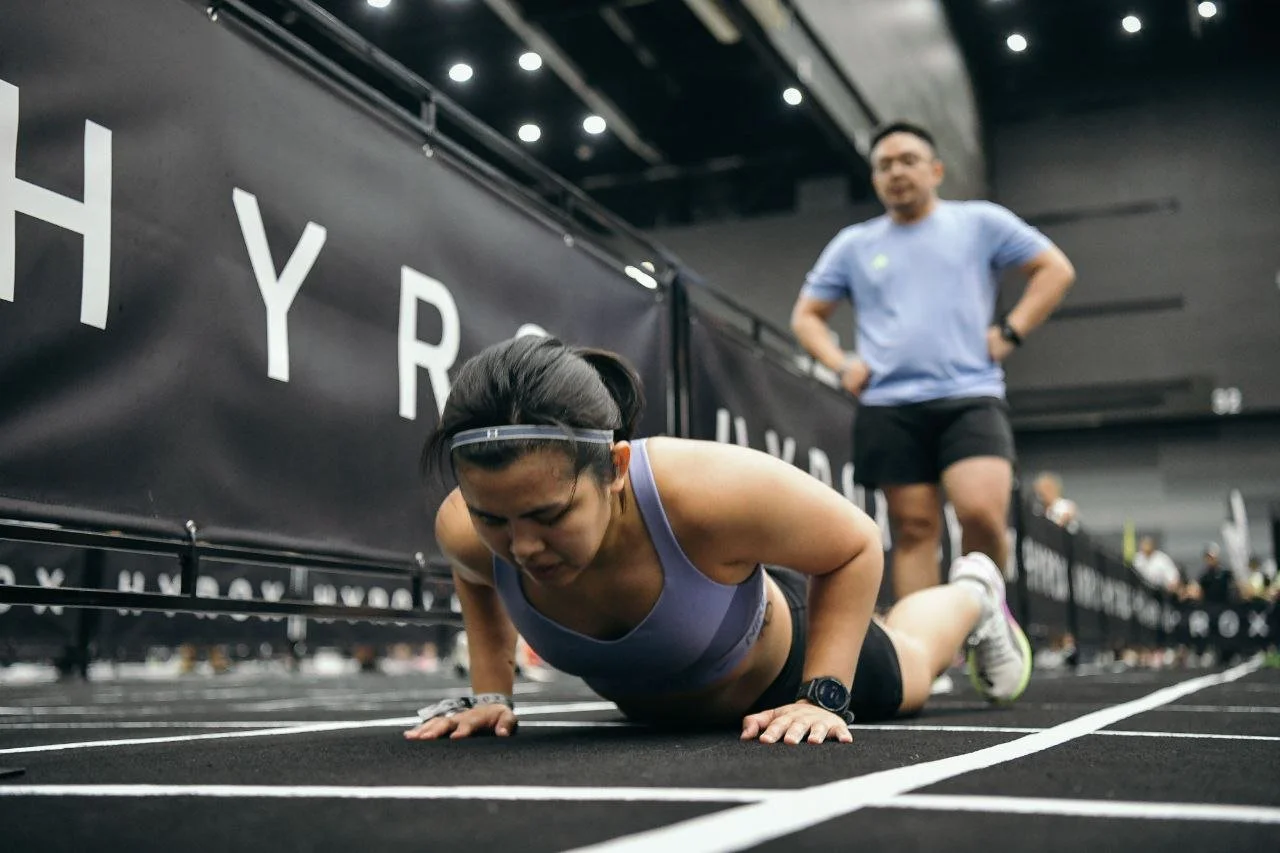 Female athlete in purple sports bra and black shorts performing a push-up on a black track at an indoor sports event, with a male observer in a blue shirt and black shorts standing nearby.
