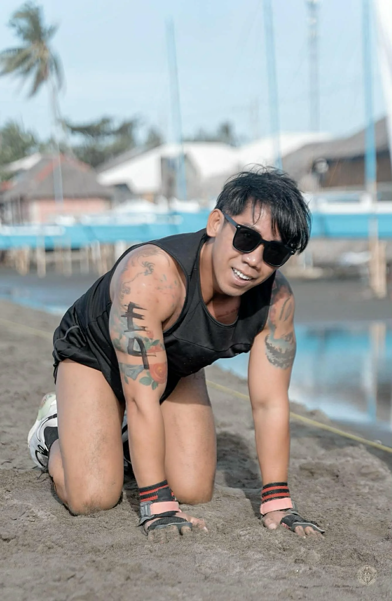 Man with tattoos on arms wearing sunglasses, black sleeveless shirt, and gloves kneeling on sandy ground with houses and sailboats in the background.