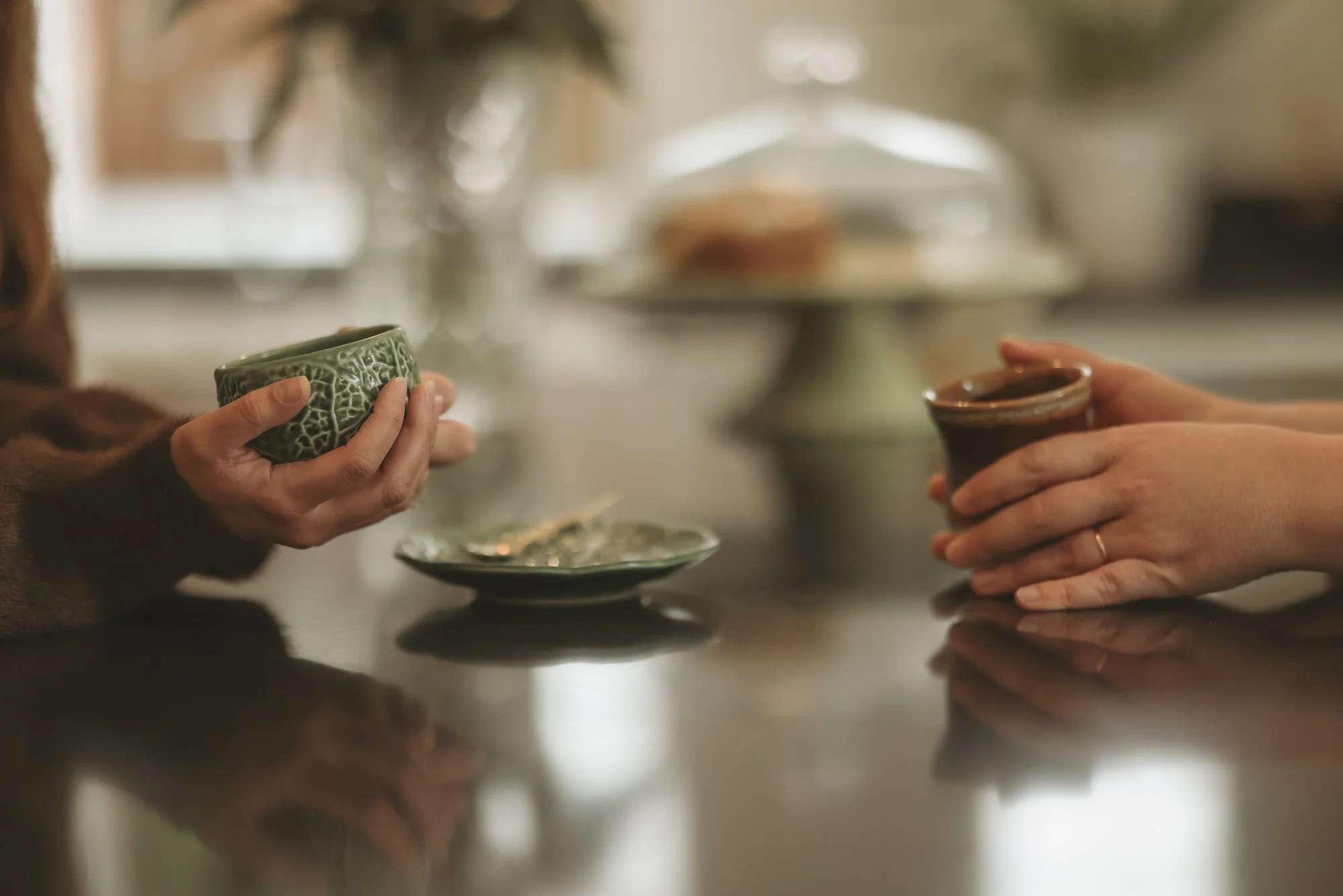 Two people holding small ceramic cups, possibly drinking tea or coffee, with a cake stand in the background on a table.