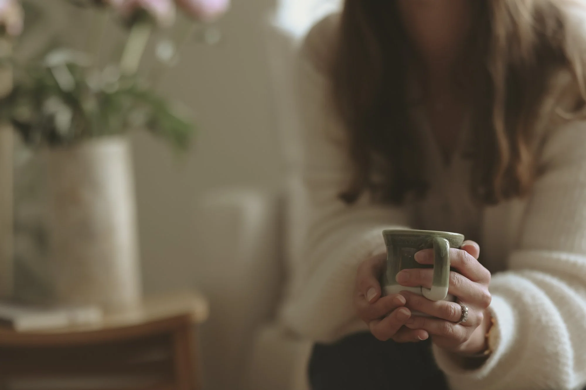 A person holding a green ceramic mug with both hands in a cozy, softly lit setting. A blurred vase with pink flowers is visible in the background.