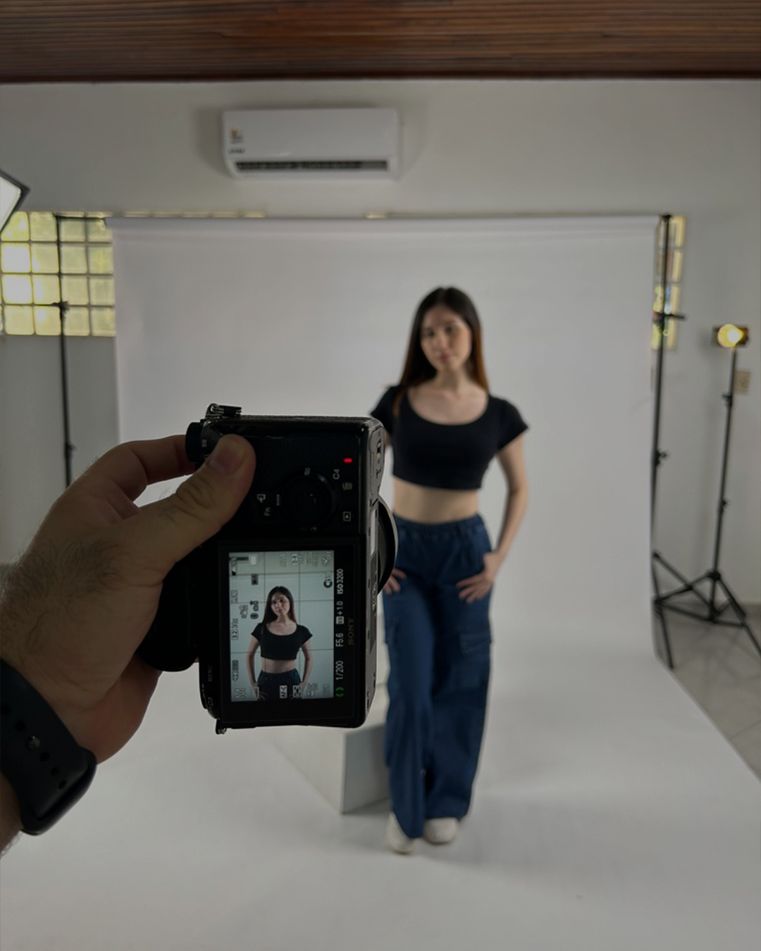 Persona sosteniendo una cámara fotográfica, enfocando a una mujer en un estudio con fondo blanco y equipo de iluminación.