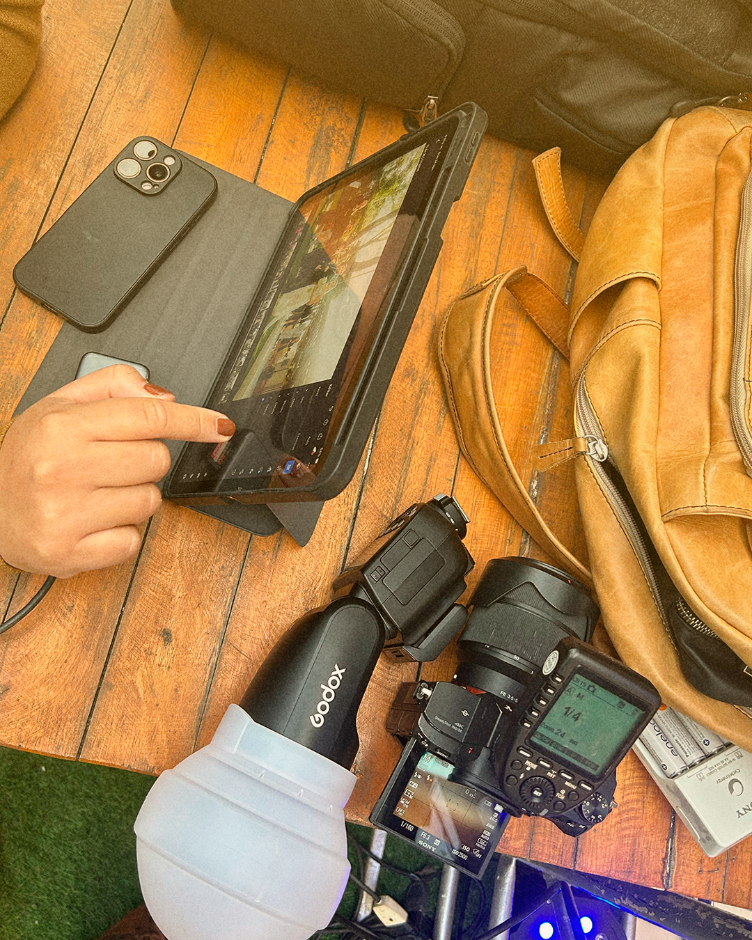 Mesa de madera con cámara, lentes, un teléfono, un drone, una bolsa de cuero y un dispositivo para fotografía, junto a una mano operando una tableta.